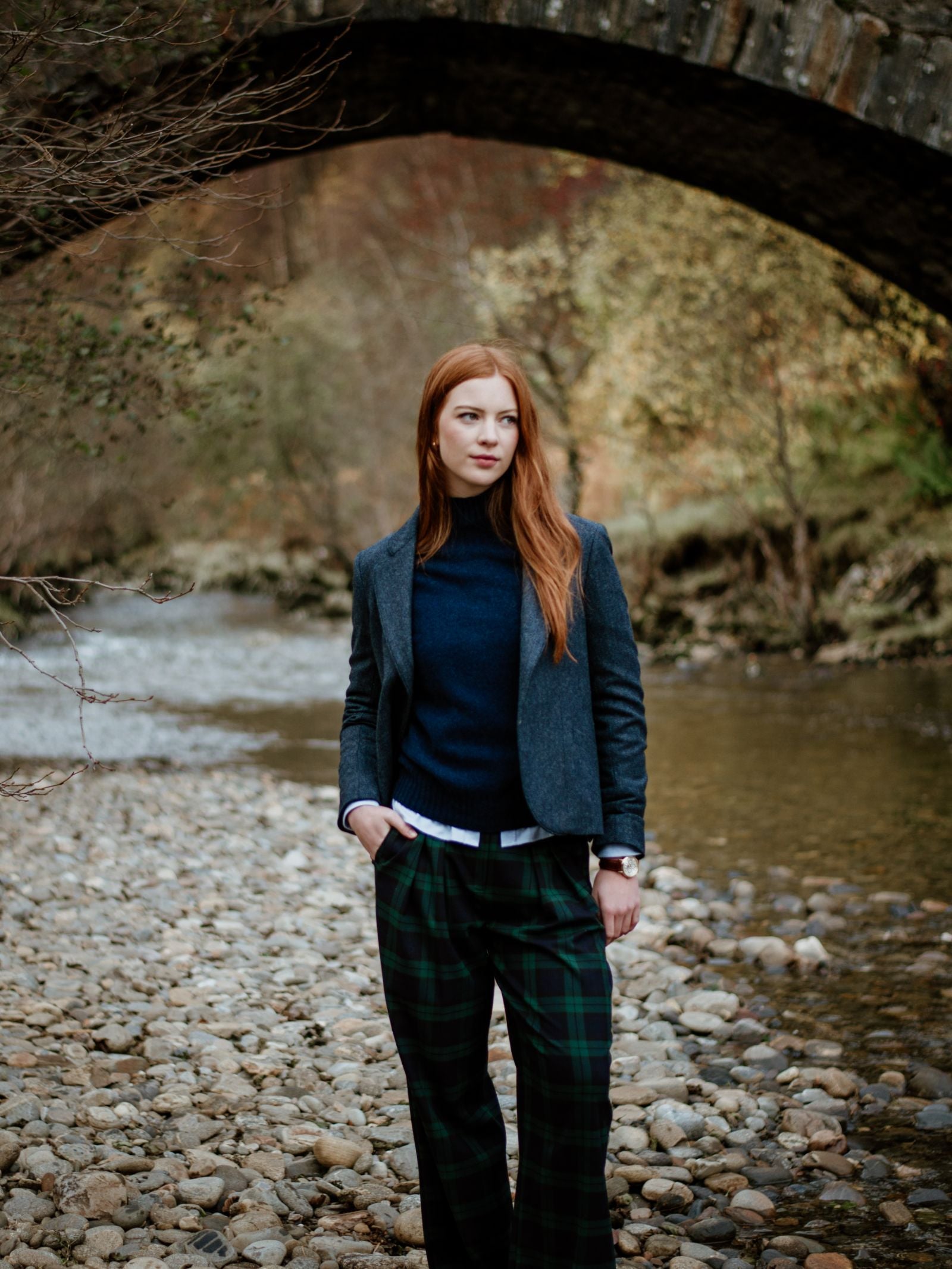Wearing the Campbell's of Beauly Auld Stock House Jacket, a woman with long red hair stands on a rocky riverbank, hands in her pockets. Behind her, an old stone bridge arches over the stream amid autumn trees.