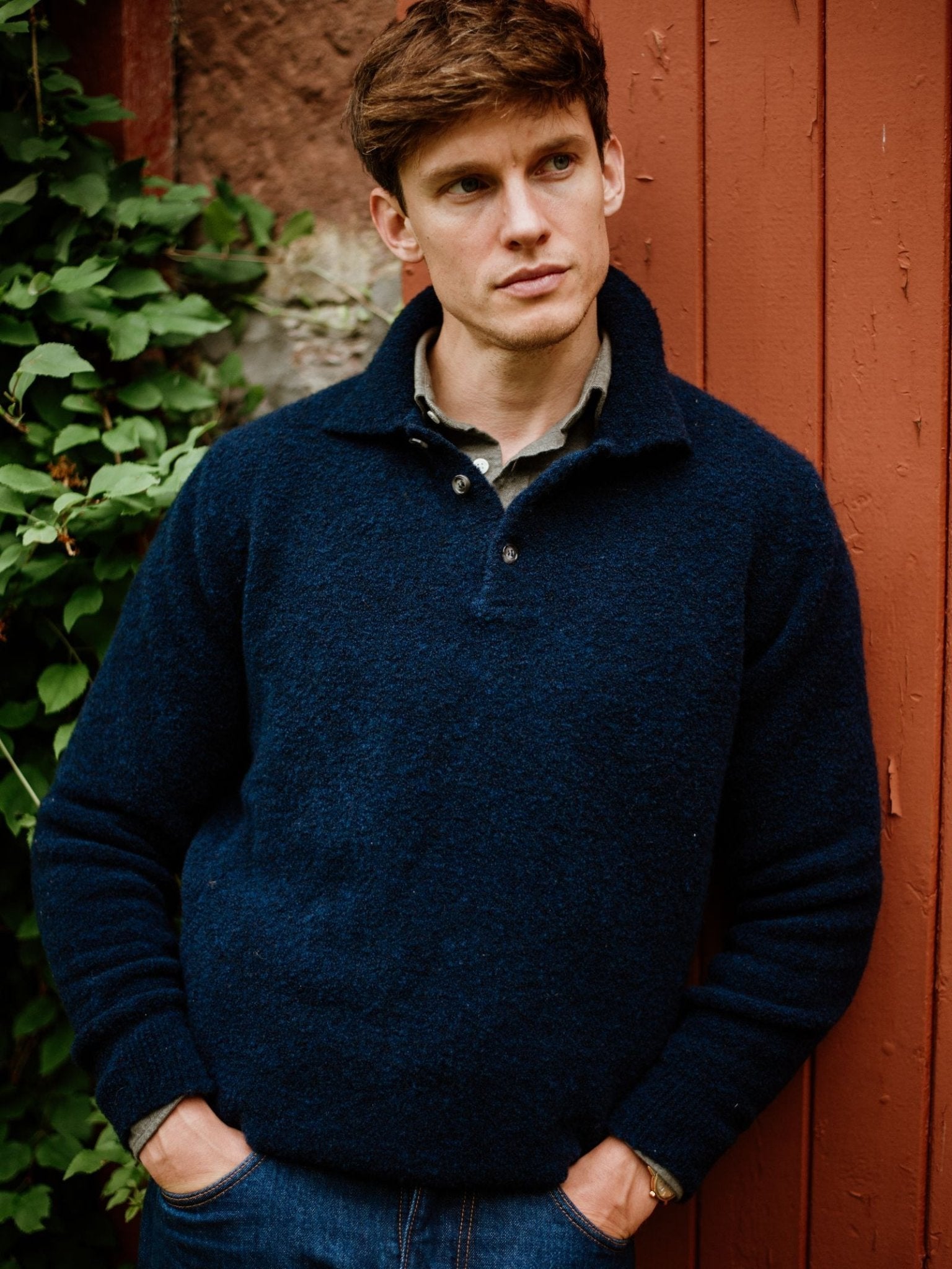 A man with short brown hair stands outdoors against a red wooden wall, wearing Campbell's of Beauly's Auld Stock Bouclé Neck Jumper and jeans, hands in his pockets and a thoughtful look. Green leaves are visible on the left.