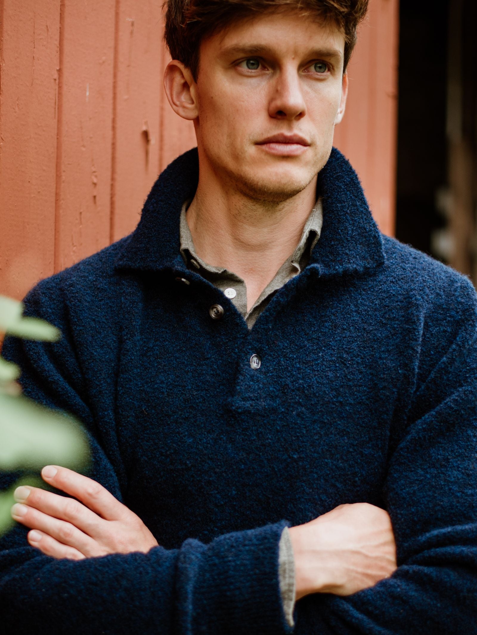 A young man with light brown hair stands before a red wooden wall, arms crossed and looking serious, wearing the Campbell's of Beauly Auld Stock Bouclé Neck Jumper over a gray shirt.
