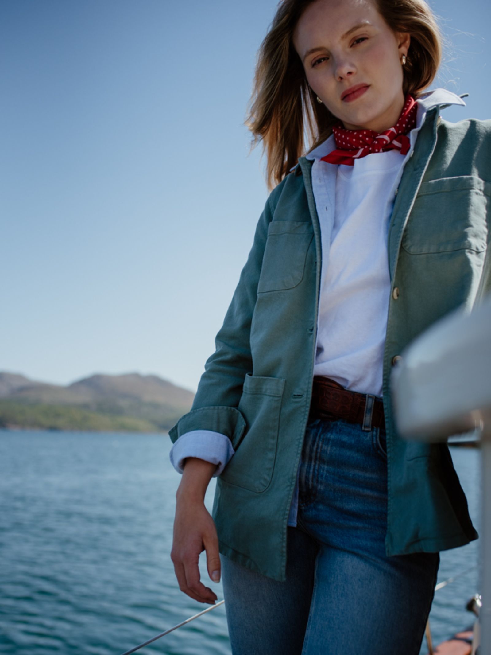 A woman stands on a boat in the sunshine, wearing the Campbell's of Beauly Auld Stock Summer Chore Jacket in green over a white shirt, red neck scarf, and jeans. Mountains and water form the scenic background.