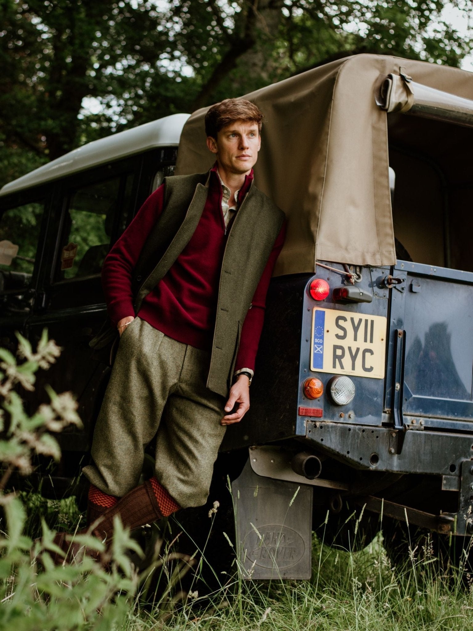 A man in country-style fieldwear leans against a vintage 4x4 in a grassy, tree-filled setting. He wears a maroon sweater, grey vest, premium wool trousers, brown boots, and Hopstitch Shooting Socks by Campbell's of Beauly.