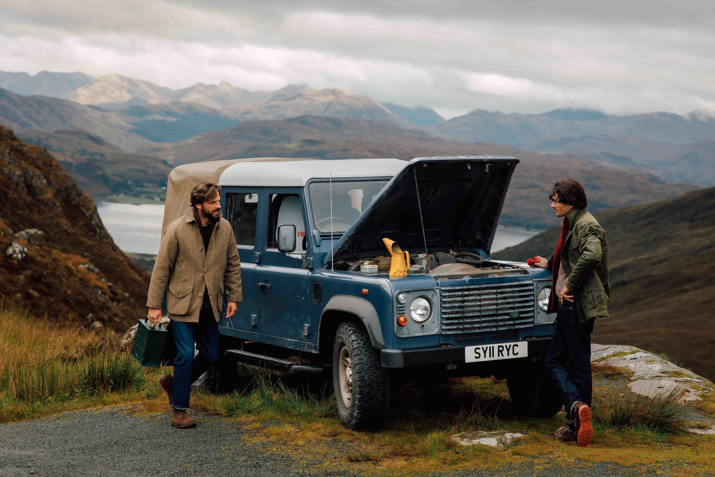 Two men stand beside a blue Land Rover with its hood open on a scenic mountain road. One man holds a green toolbox; mountains and a lake are visible in the background under a cloudy sky.