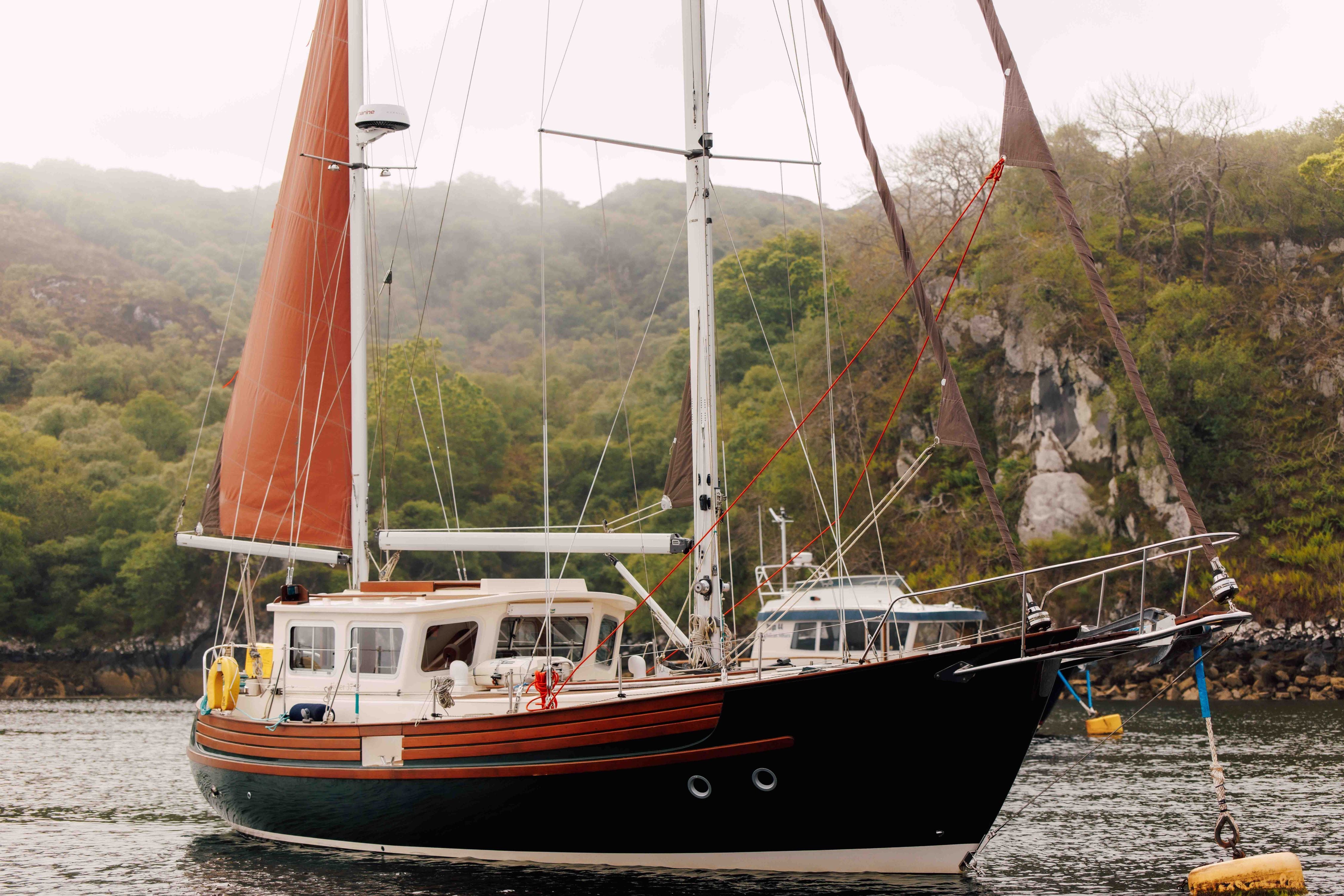 A black and brown sailboat with tan sails is anchored on calm water near a forested shoreline and rocky cliffs, with another boat partially visible in the background.