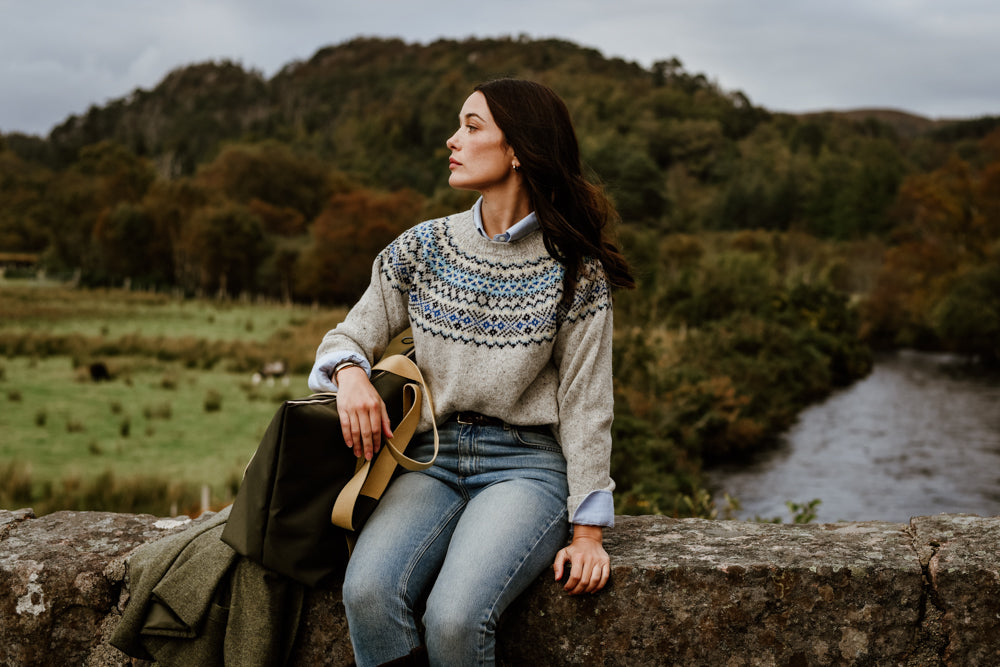 A woman in a patterned sweater and jeans sits on a stone wall, holding a bag, with a river and green hills in the background under a cloudy sky.