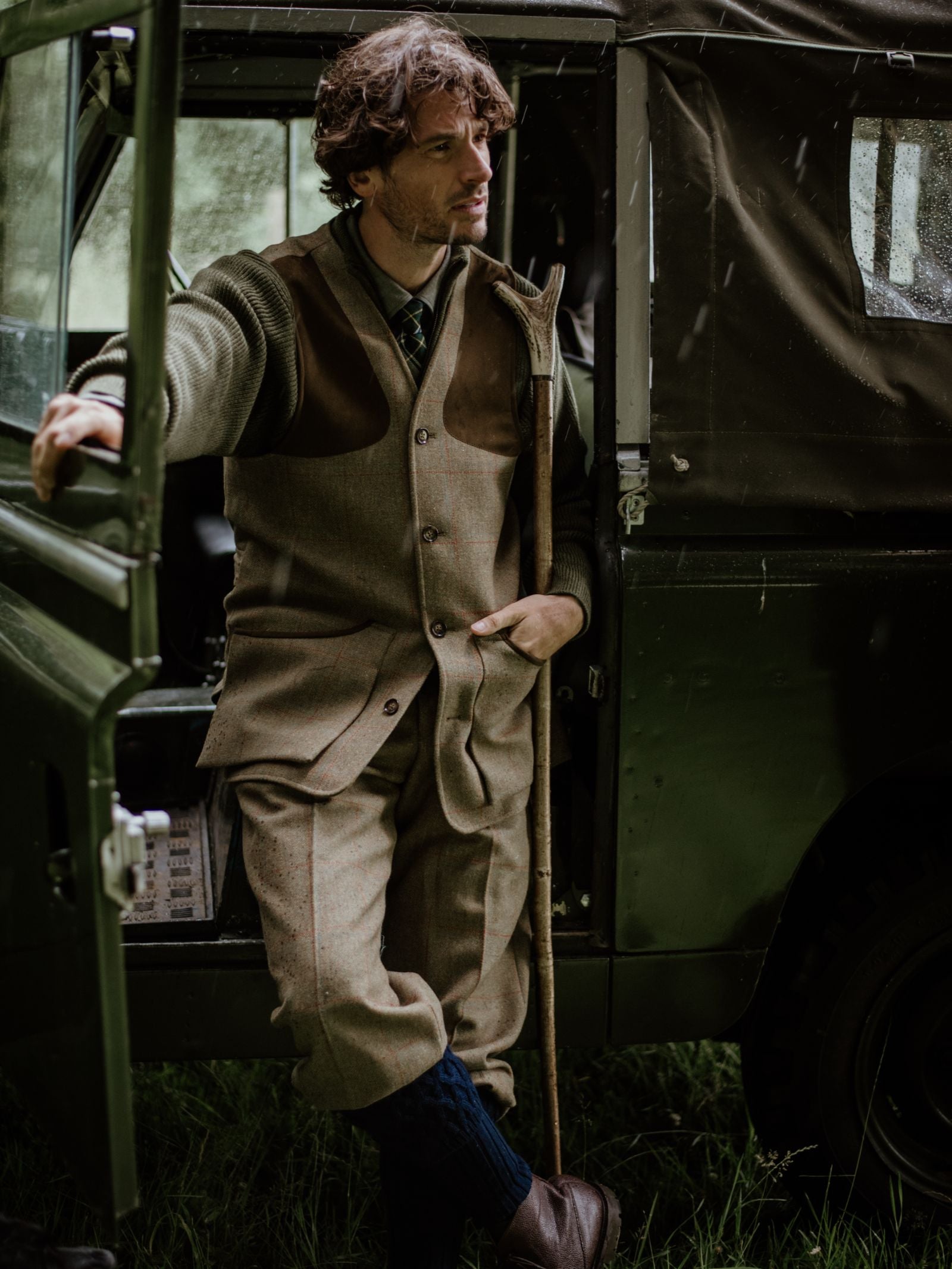 A man stands by an open jeep door in the countryside, wearing Campbell's of Beauly Tweed Shooting Waistcoat House Tweed, with matching trousers and knee-high socks; greenery and a dark vehicle are visible behind him.