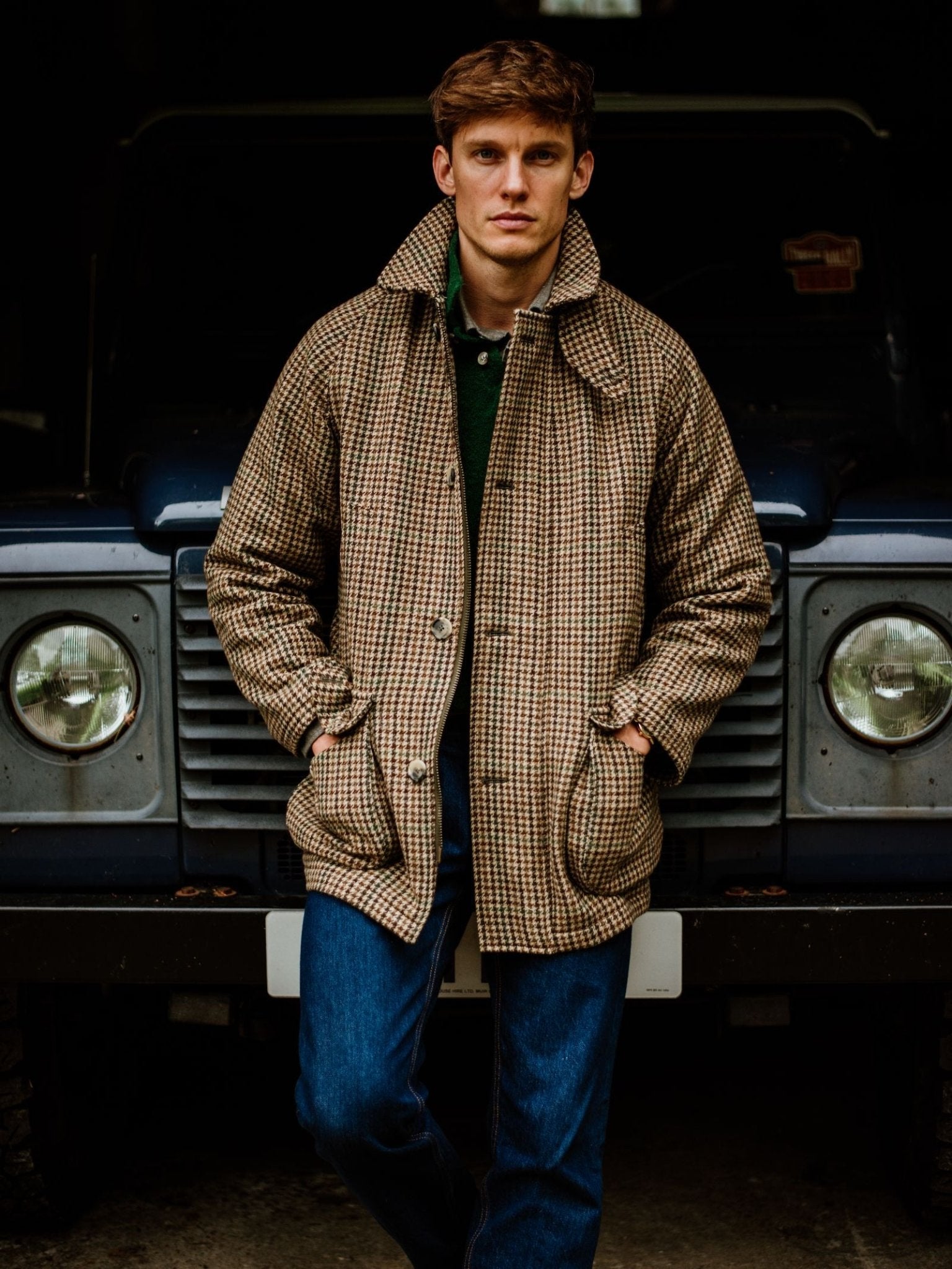 A man wearing the Cannich Coat by Campbell's of Beauly and blue jeans stands in front of a dark vintage SUV, hands in his pockets, looking directly at the camera.