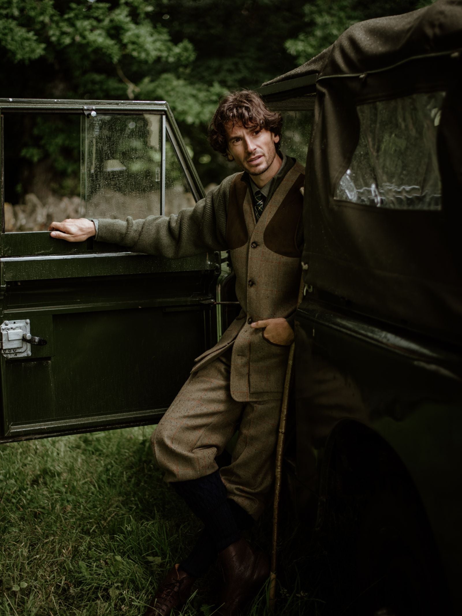 A man in vintage-style brown tweed and the Campbell's of Beauly Tweed Shooting Waistcoat House Tweed leans against an old dark green vehicle with the door open, set on grass with trees behind, evoking classic countryside pursuits.