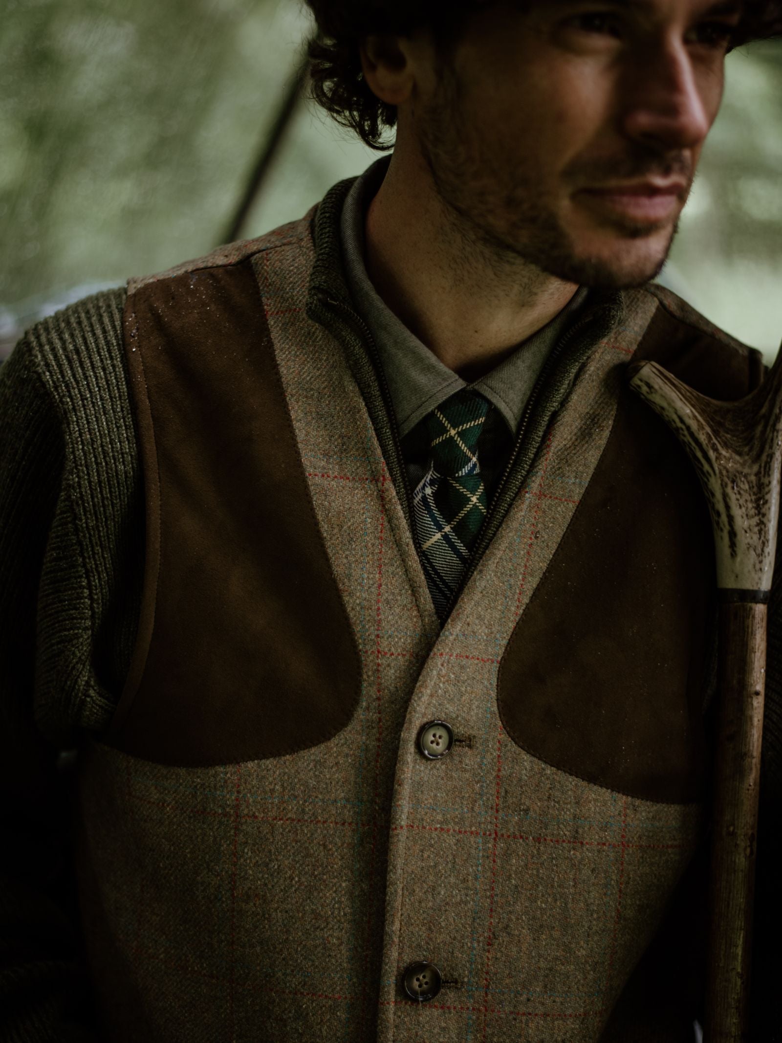 A man in a Campbell's of Beauly Tweed Shooting Waistcoat House Tweed, paired with a green plaid tie and shirt, holds an antler. Soft lighting and his partially visible face evoke a rustic, countryside feel.