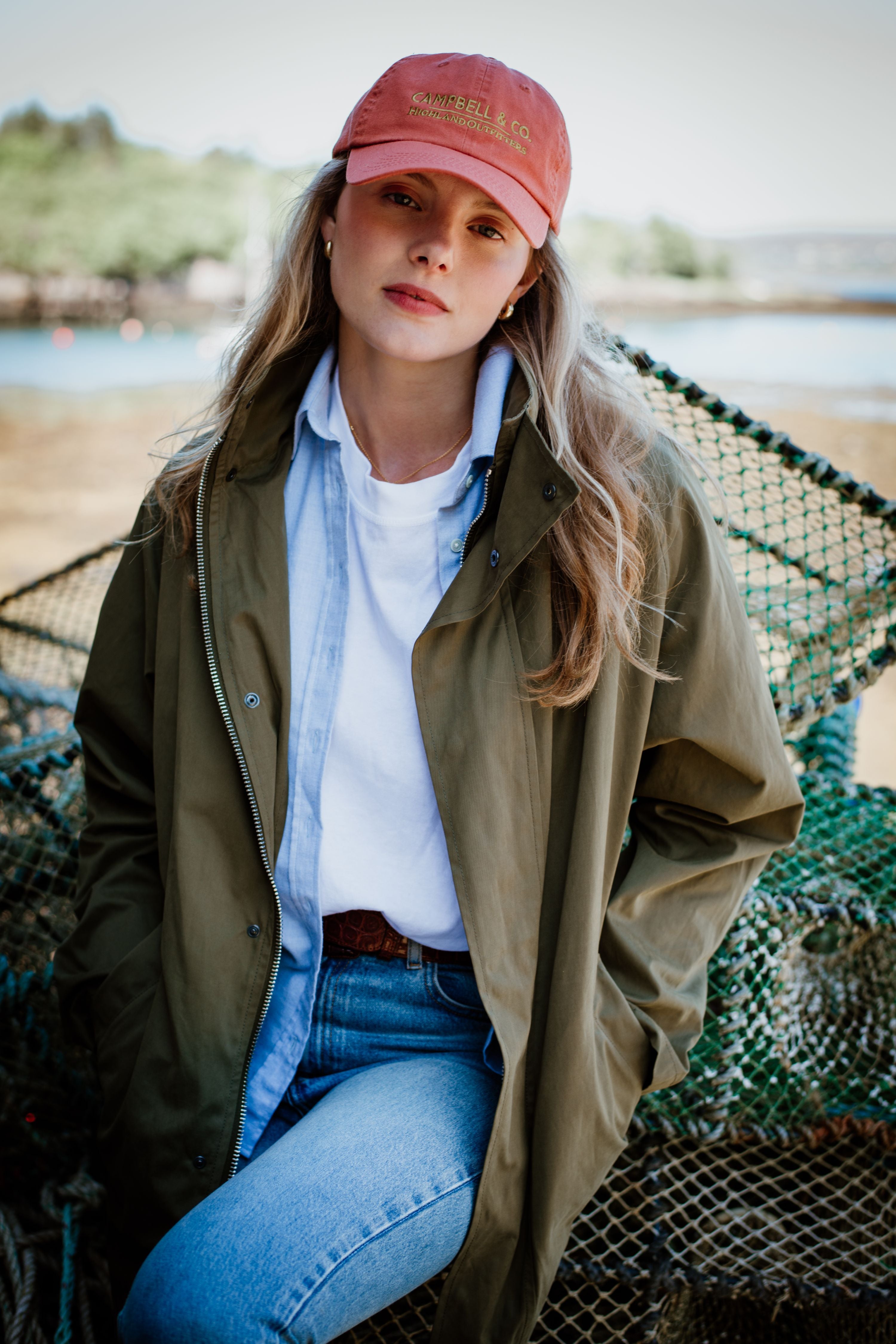A woman with long blonde hair wears a red cap, olive-green jacket, white t-shirt, and jeans. She stands outdoors near stacked fishing nets, looking at the camera with a neutral expression.