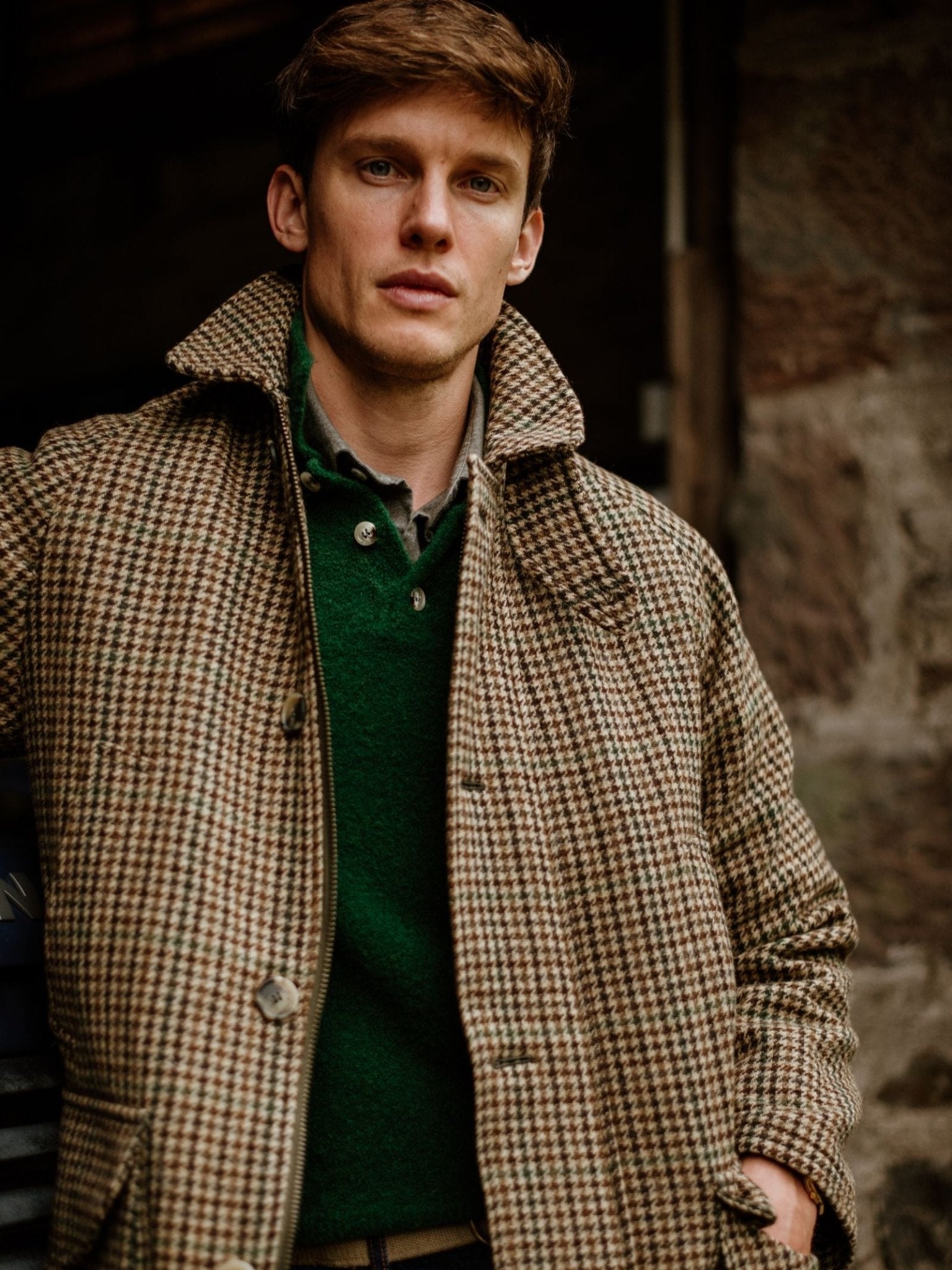 A man with light brown hair wears the Cannich Coat by Campbell's of Beauly over a dark green sweater, standing before a stone wall and gazing calmly at the camera.