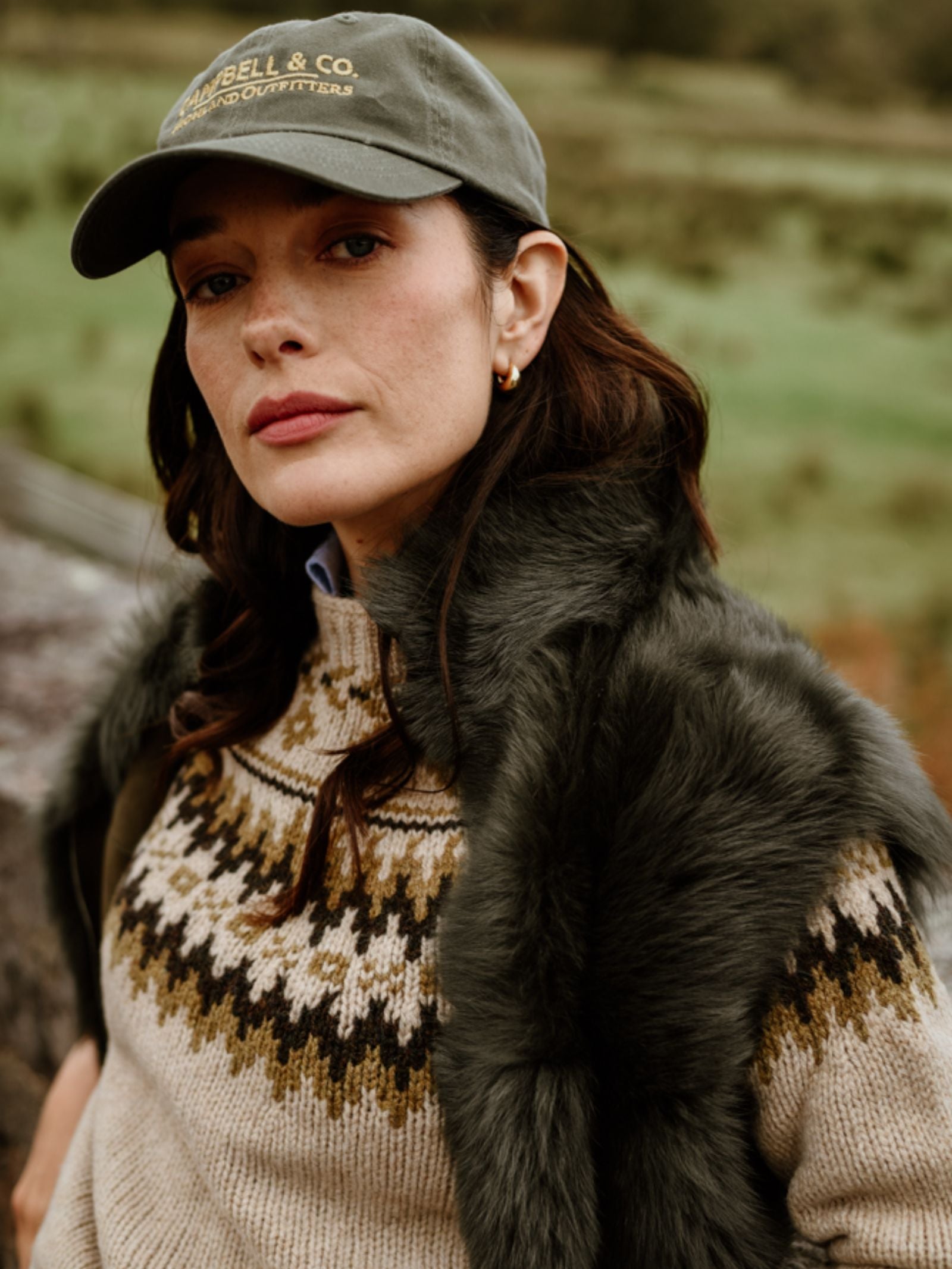 A woman with dark hair and a calm expression stands outdoors against a blurred grassy landscape, wearing the Campbell's of Beauly Auld Stock Sheepskin Reversible Gilet over a patterned knit sweater and a green baseball cap.