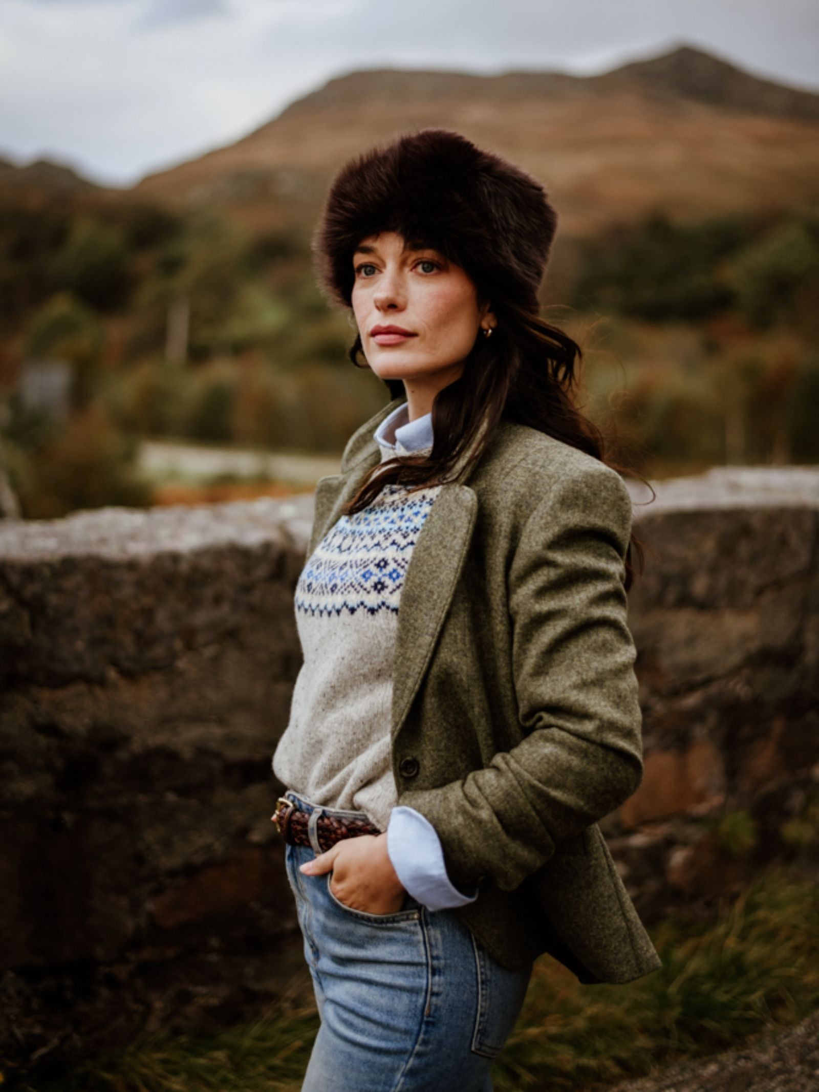 A woman wearing the Campbell's of Beauly Pillar Box Hat, green blazer, patterned sweater, and jeans stands outdoors by a stone wall with hills and trees in the background on a cloudy day.