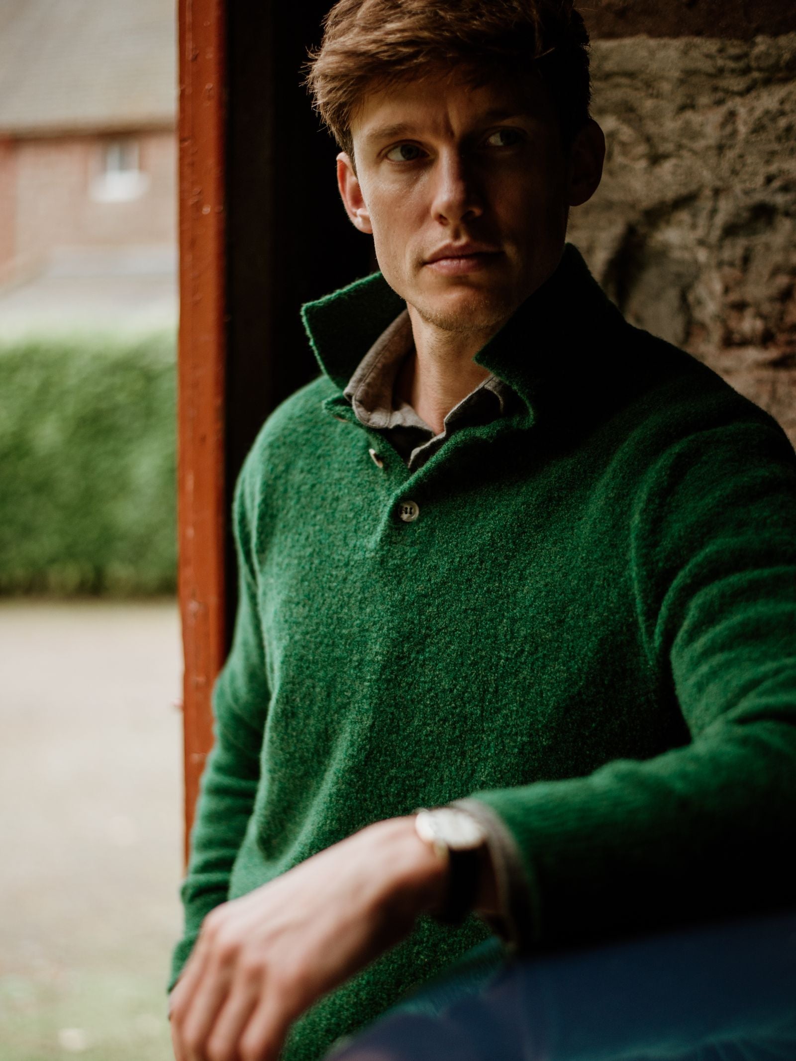 A man with light brown hair stands indoors near a doorway, gazing thoughtfully aside, wearing the Campbell's of Beauly Auld Stock Bouclé Neck Jumper in textured green. A garden and building are visible outside through a window.