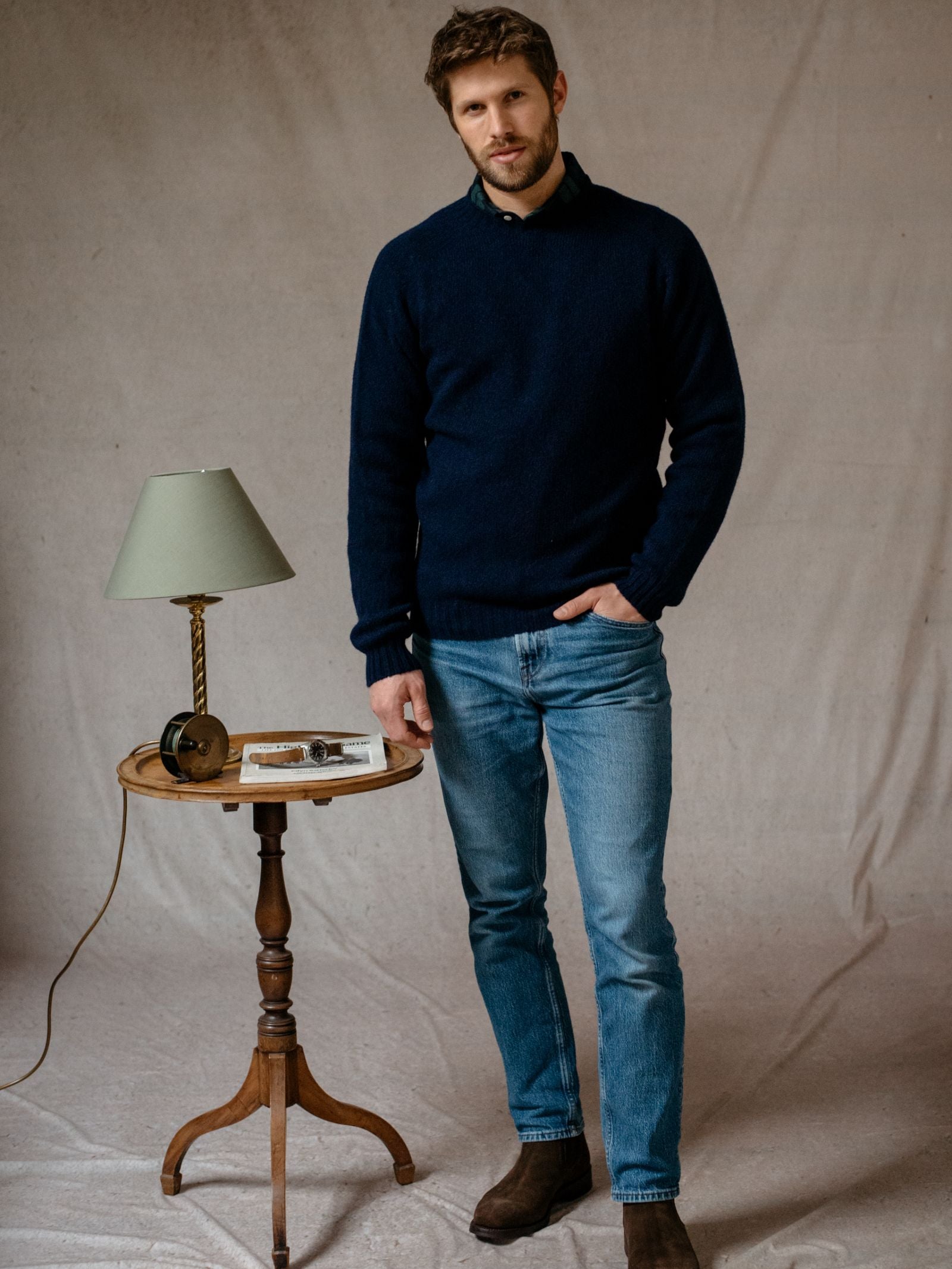 A man with short brown hair wears a navy Shetland Jumper by Campbell's of Beauly, light blue jeans, and brown boots. He stands indoors beside a small wooden table with a lamp, book, and glasses on it against a plain beige fabric background.