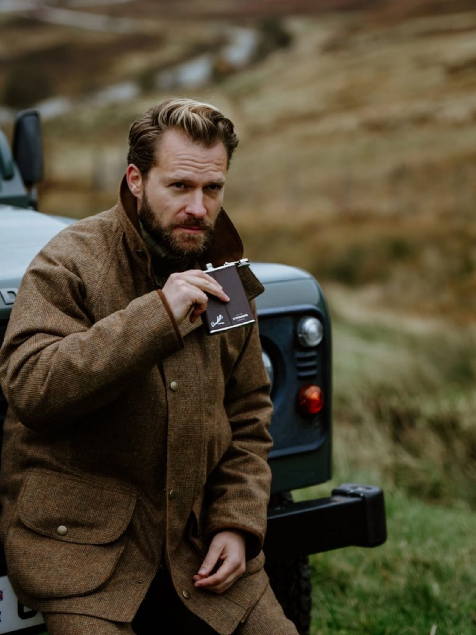 A bearded man in a brown tweed jacket sits on a green vehicle, holding the Campbell's of Beauly x Ettinger 6oz Chocolate Hip Flask, with a rugged grassy landscape in the background.