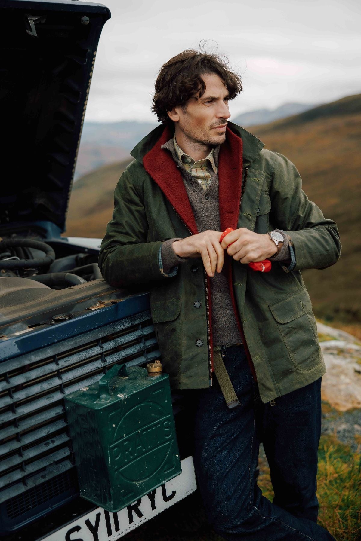 A man in an olive green jacket leans against a blue SUV with the hood open, holding a red cloth. Hills and a cloudy sky are visible in the background, suggesting a rural or remote location.