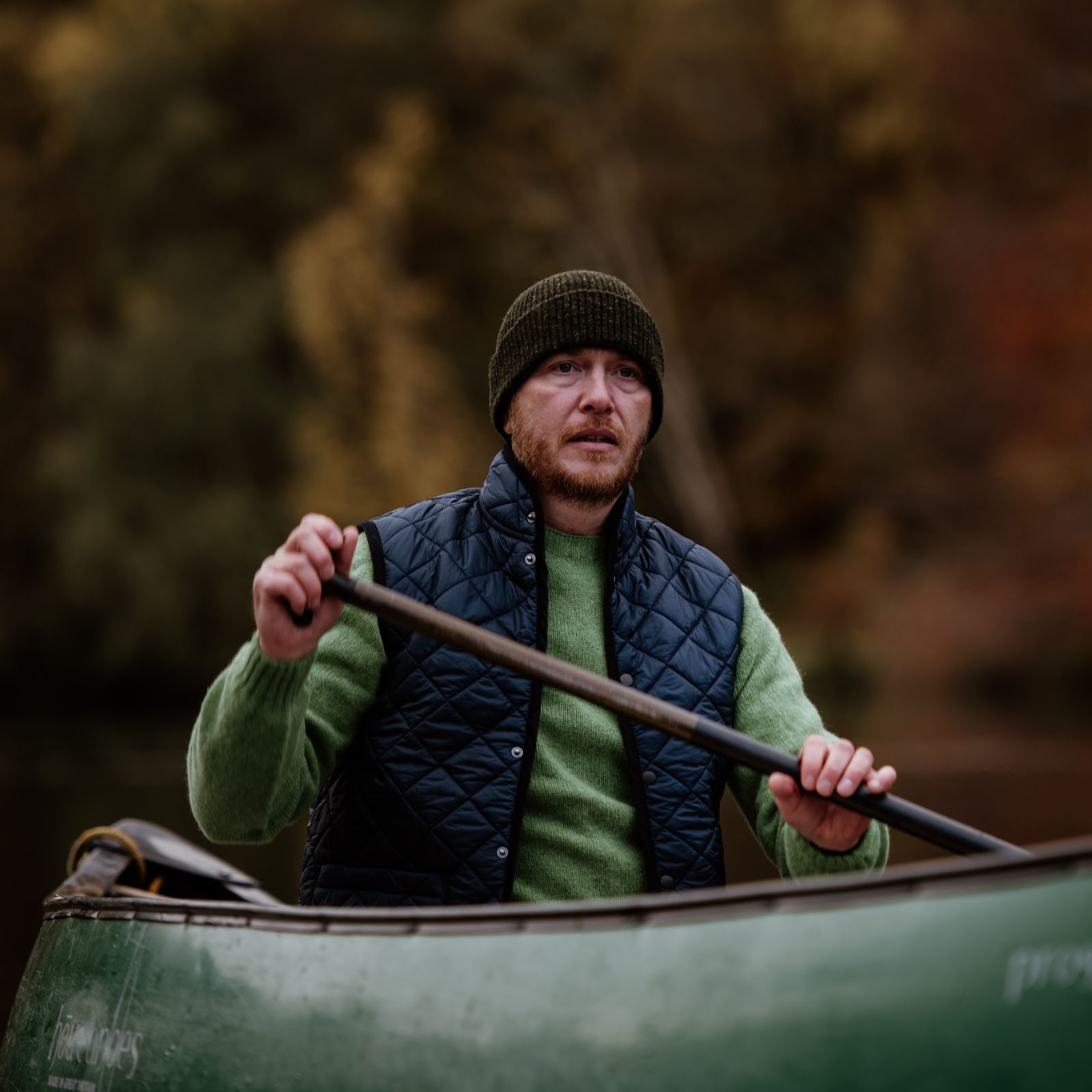 A man wearing a green sweater, black quilted vest, and green knit hat paddles a green canoe on a calm body of water with autumn-colored trees in the background.