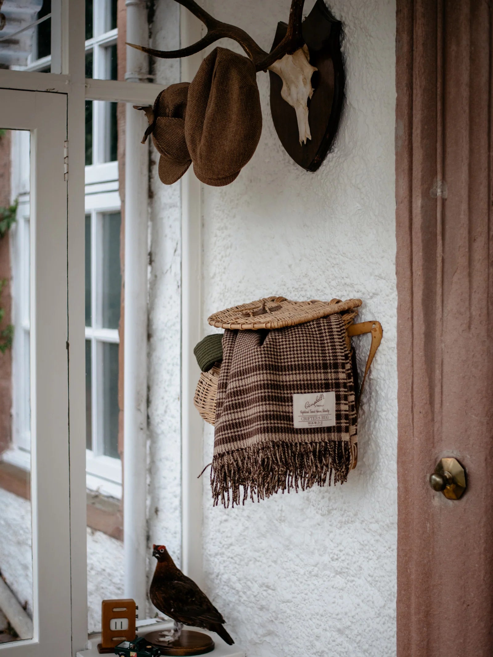 A Crofters Blanket from Campbell's of Beauly brings warmth to a country-style entryway, where hats and a scarf hang below mounted antlers, with a small table displaying a bird figurine, key holder, and more for added charm.