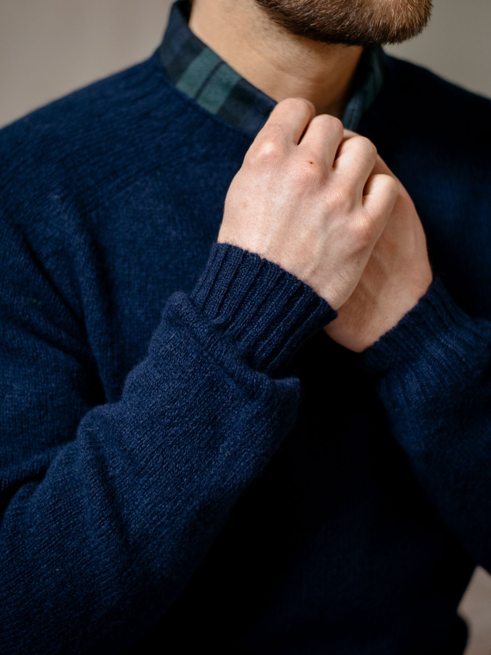 A man adjusts the collar of a Campbell's of Beauly Tartan Shirt under a navy sweater, with only his hands, torso, and part of his bearded face visible.