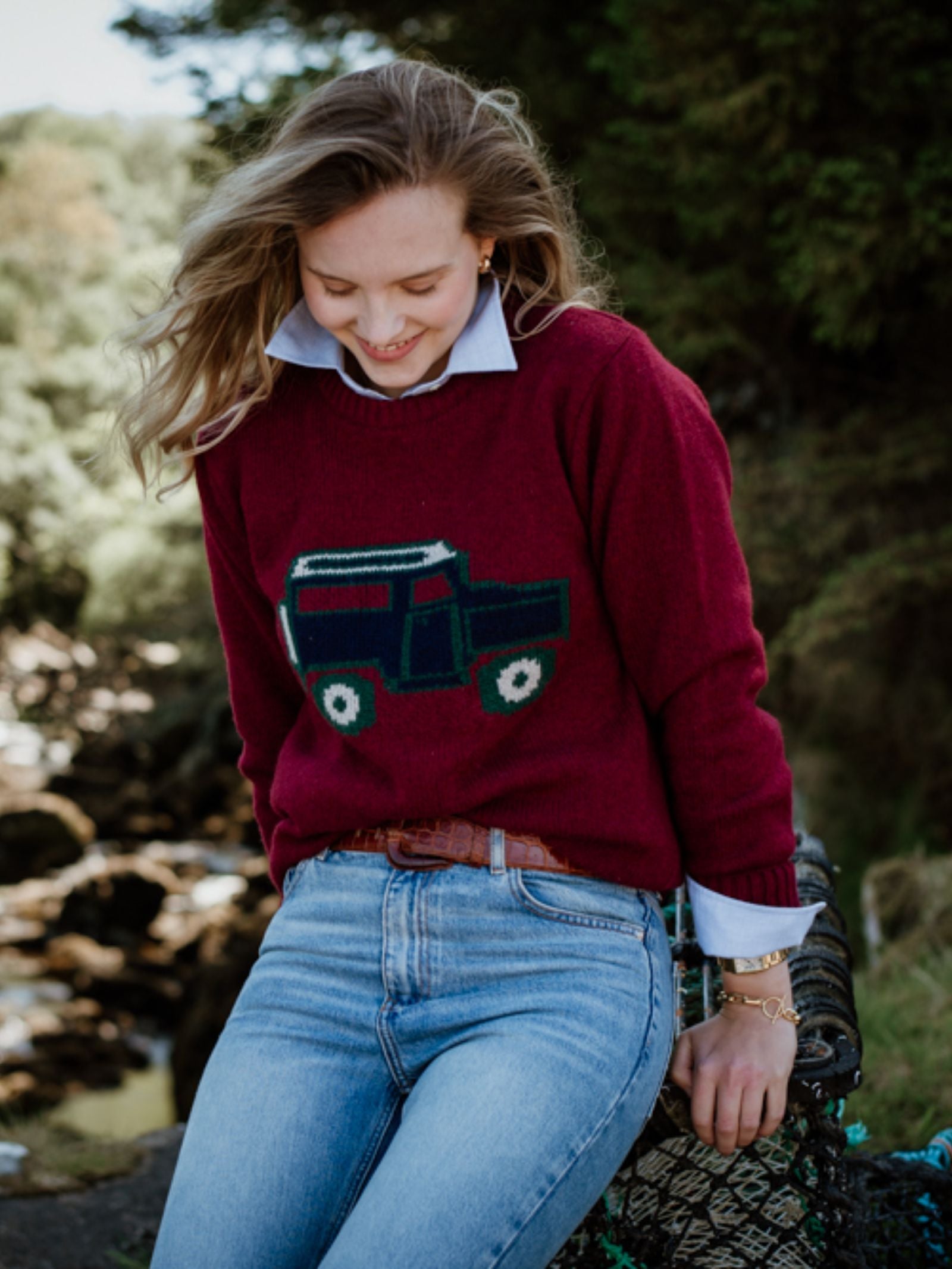 A woman with light brown hair smiles outdoors, wearing a Campbell's of Beauly Unisex Land Rover Defender Jumper in maroon over a white collared shirt and blue jeans. Trees and rocks in the background evoke British motoring charm.