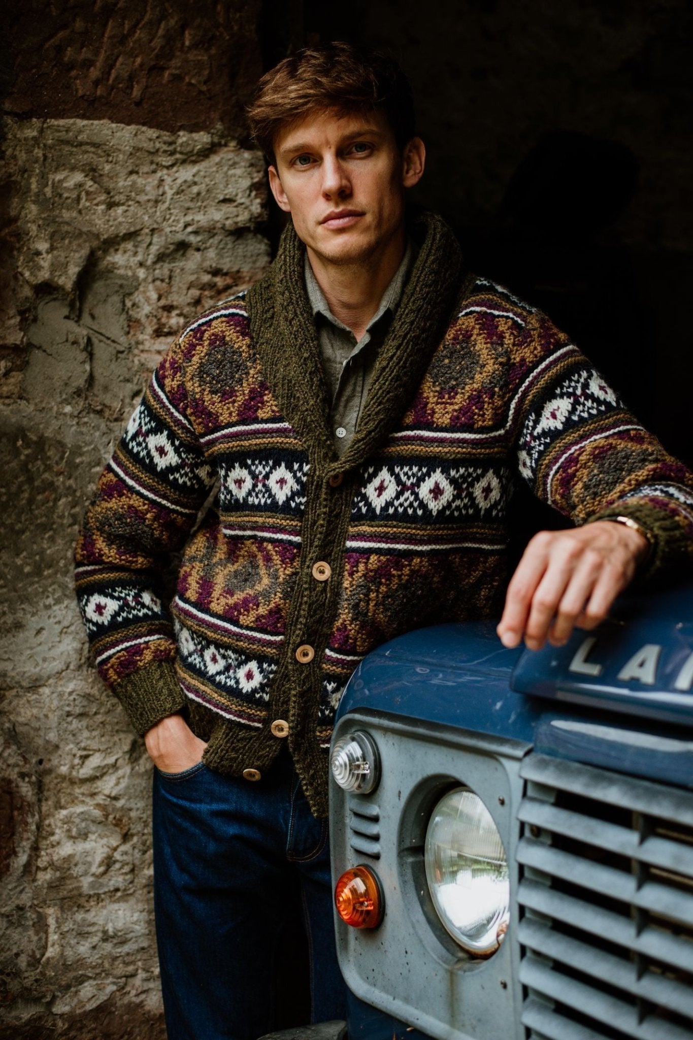 A man in a Campbell's of Beauly Navajo Shawl Collar Cardigan leans against a vintage blue Land Rover by a stone wall, looking seriously at the camera.
