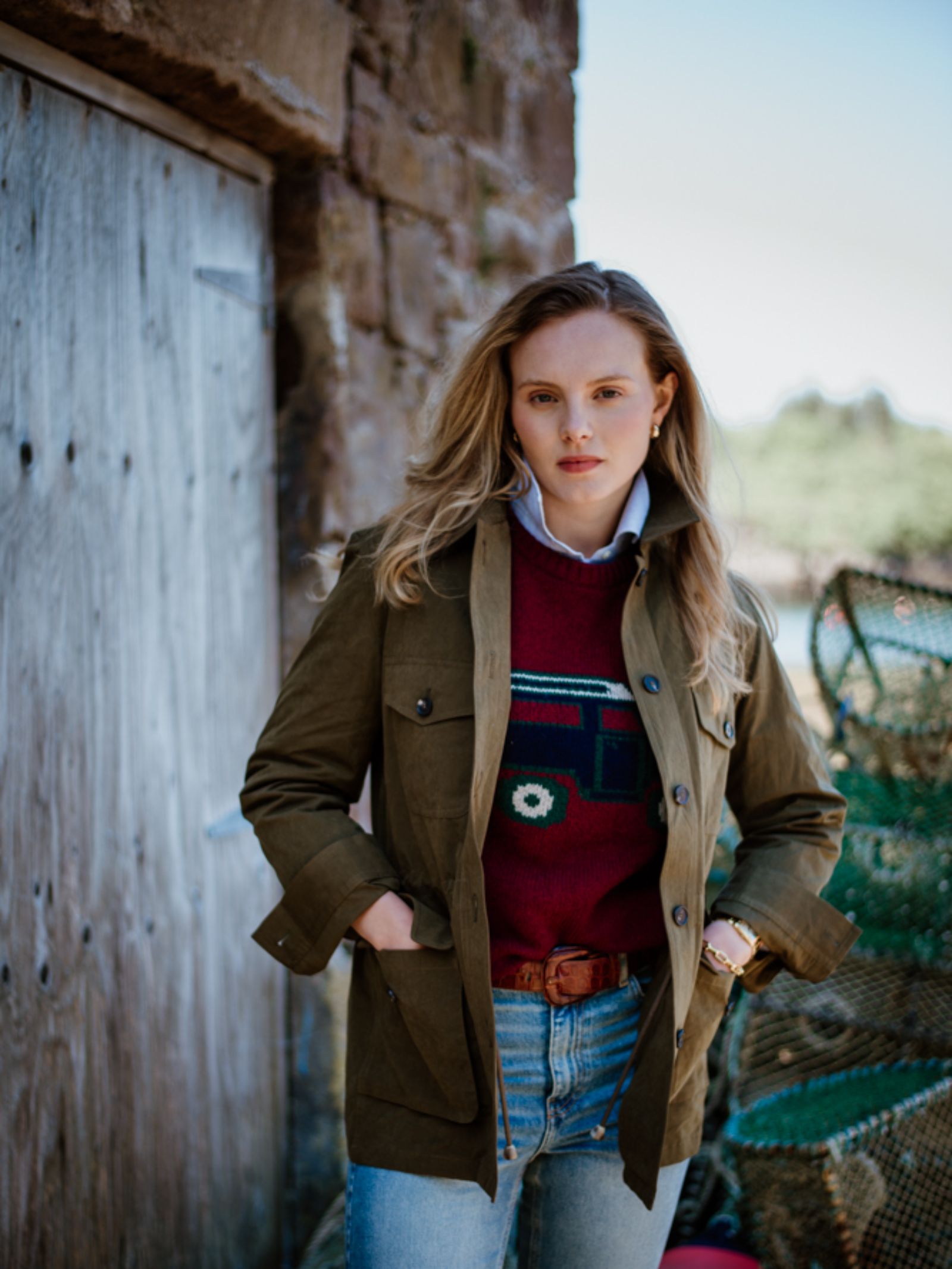 A young blonde woman wears the Campbell's of Beauly Auld Stock Aigas Field Jacket, crafted from water-repellent peach skin fabric, over a maroon graphic sweater and blue jeans, standing with hands in pockets before a wooden door and stone wall.