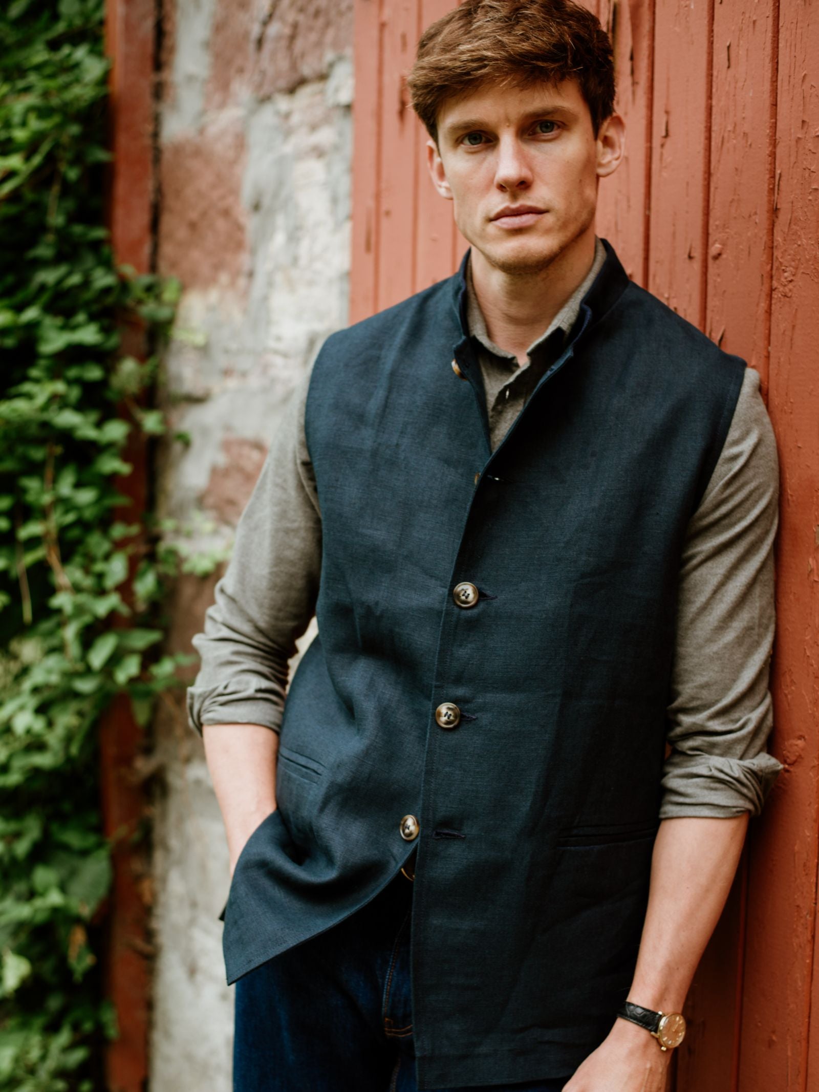 A young man with light brown hair stands against a rustic red wooden wall, wearing a gray shirt under the Linen Nehru Gilet by Campbell's of Beauly—the ideal summer layer. He looks at the camera with a neutral expression, one hand in his pocket.