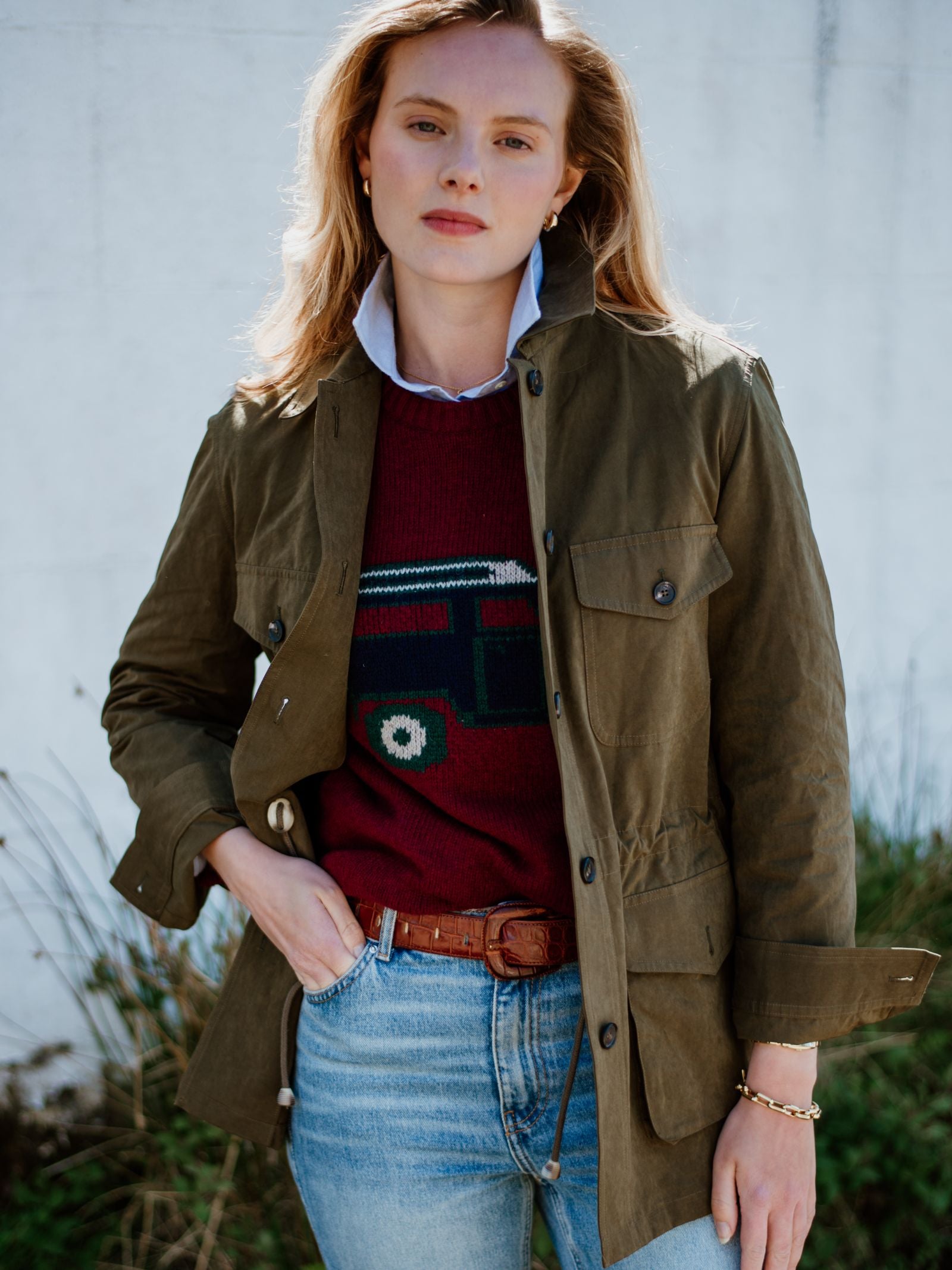 A woman with blonde hair wears the Campbell's of Beauly Auld Stock Aigas Field Jacket, made from water-repellent peach skin fabric, over a maroon sweater with a vehicle design, a blue collared shirt, and blue jeans outdoors by a white wall and plants.