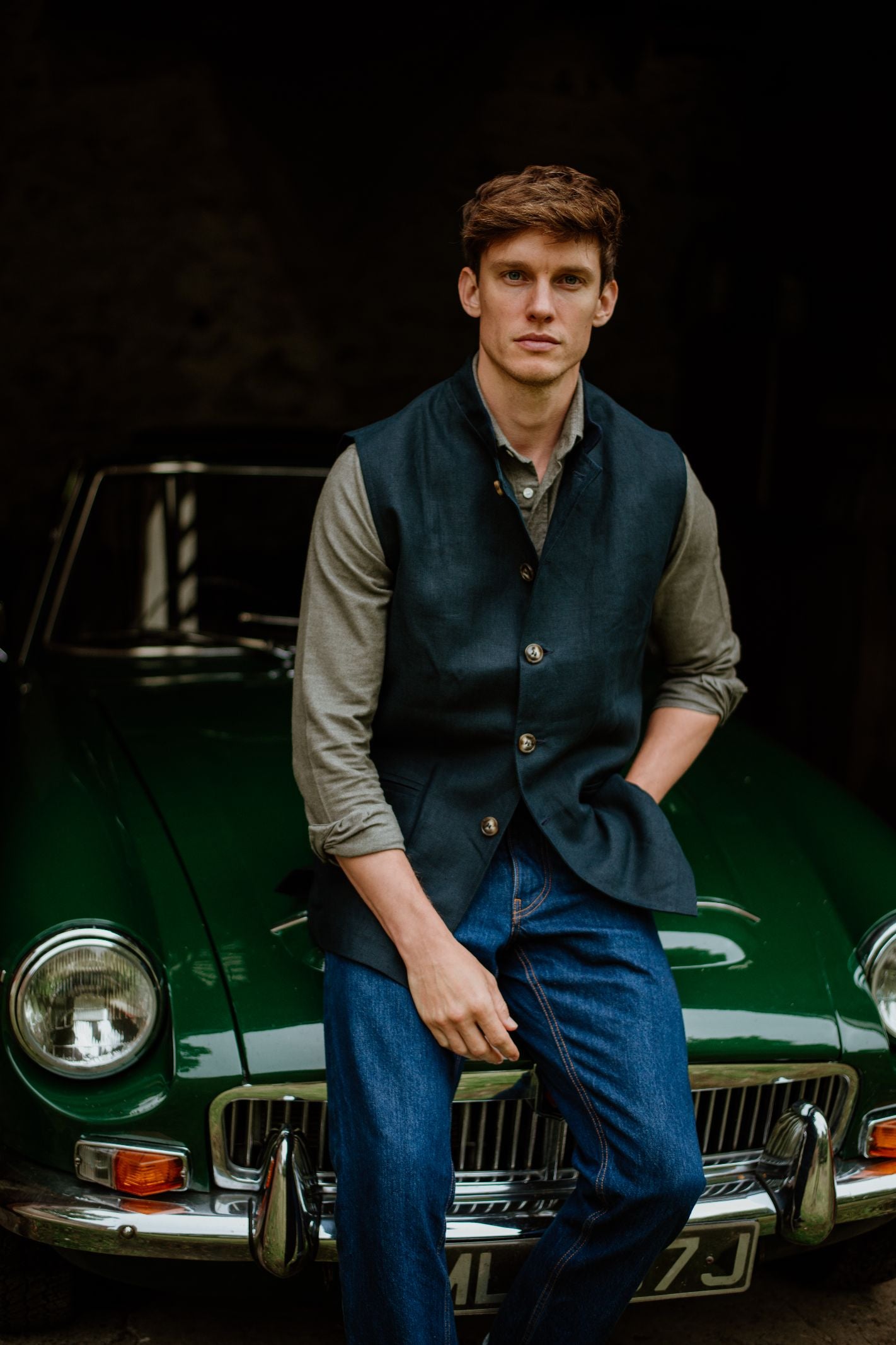 A young man with short brown hair, in a gray shirt, blue jeans, and Campbell's of Beauly Linen Nehru Gilet, sits on the hood of a classic green car, looking directly at the camera with a neutral expression.