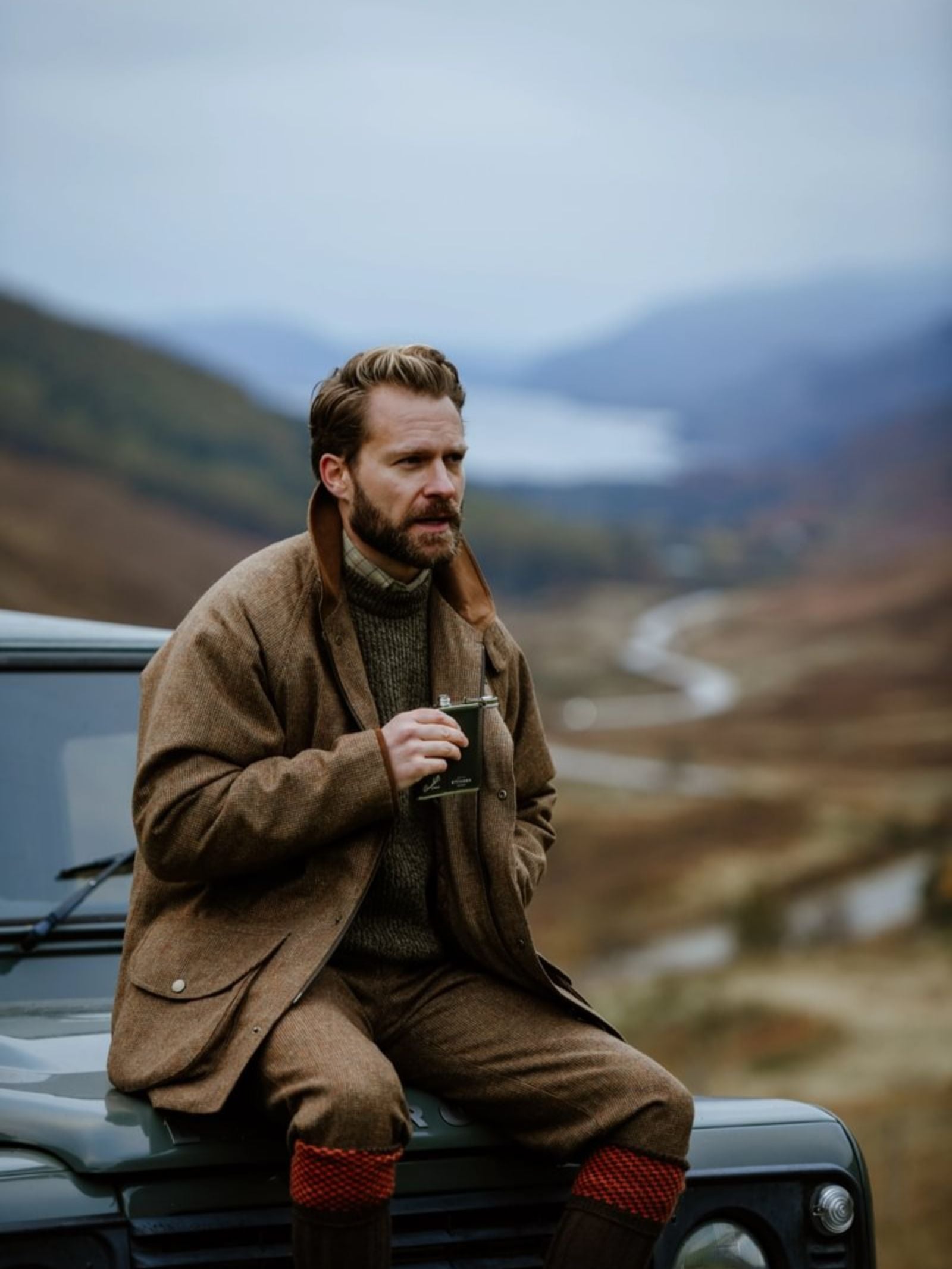 A bearded man in brown outdoor clothing holds a Campbell's of Beauly x Ettinger 6oz Forest Green Hip Flask while sitting on a green vehicle, with hills and a winding river behind him under an overcast sky.