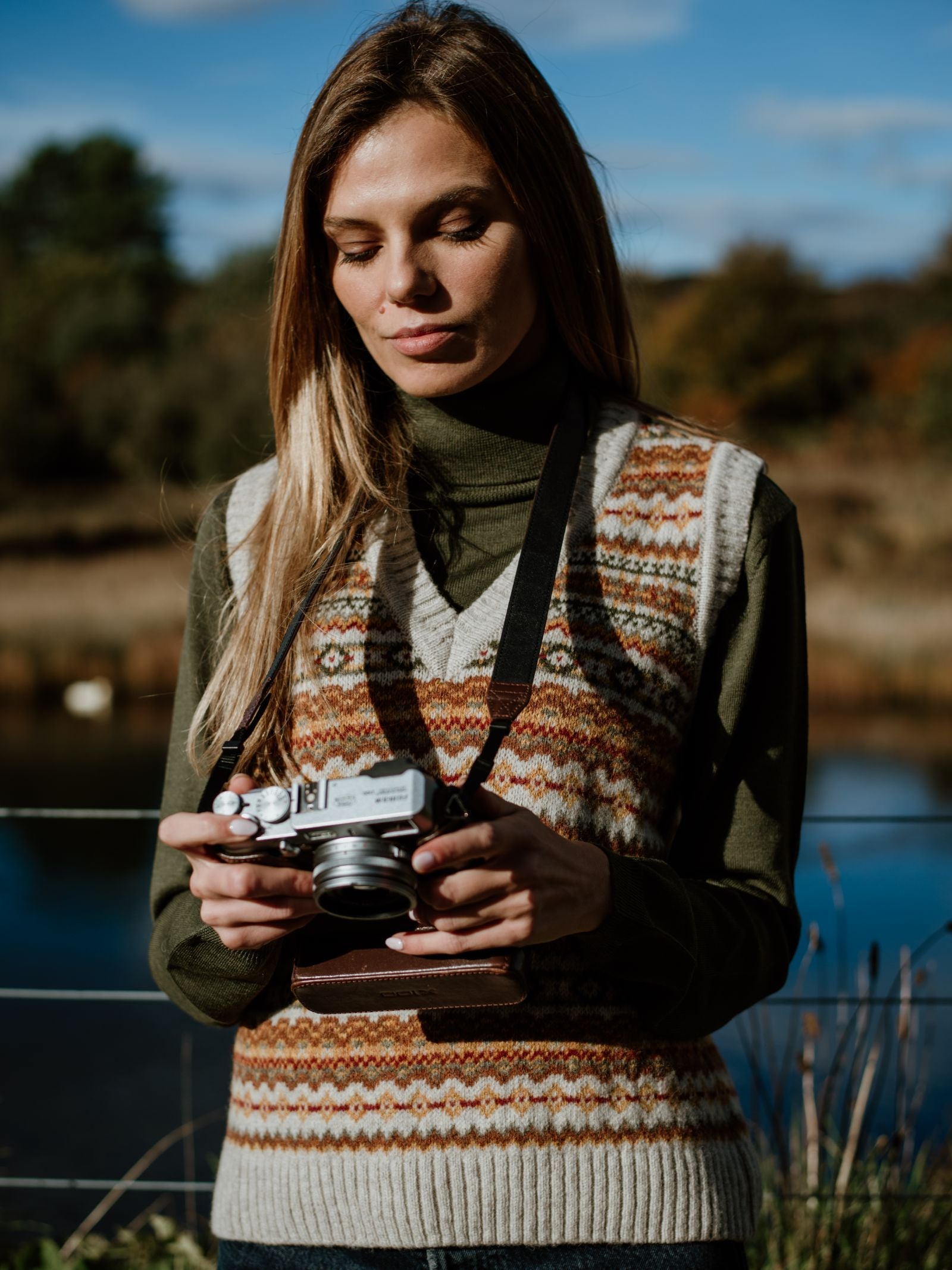 A woman with long blond hair stands outdoors holding a vintage camera, wearing Campbell's of Beauly's Auld Stock Fairisle V-Neck Tank over a green turtleneck. A blurred landscape of water, trees, and Scotland’s blue sky forms the backdrop.