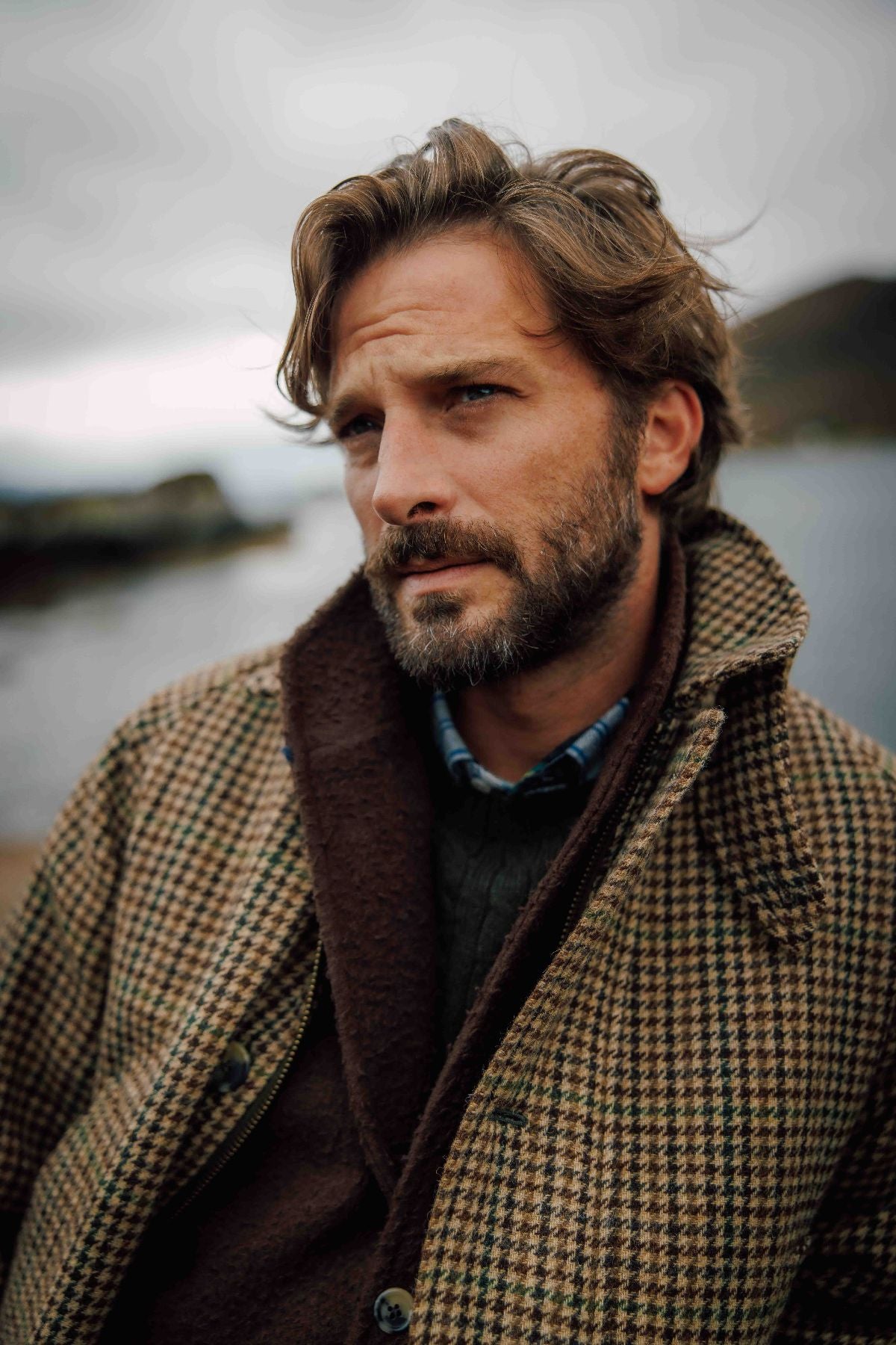 A man with wavy brown hair and a beard looks thoughtfully into the distance, wearing a brown checked coat over a sweater and shirt, standing outdoors near water on a cloudy day.