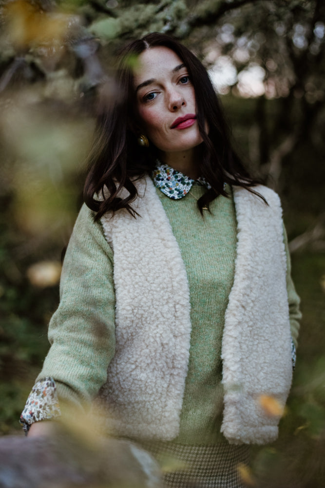 A woman with long dark hair stands outdoors in a wooded area, wearing a light green sweater, a floral collared shirt, and a cream-colored sherpa vest. She looks thoughtfully at the camera with a soft expression.