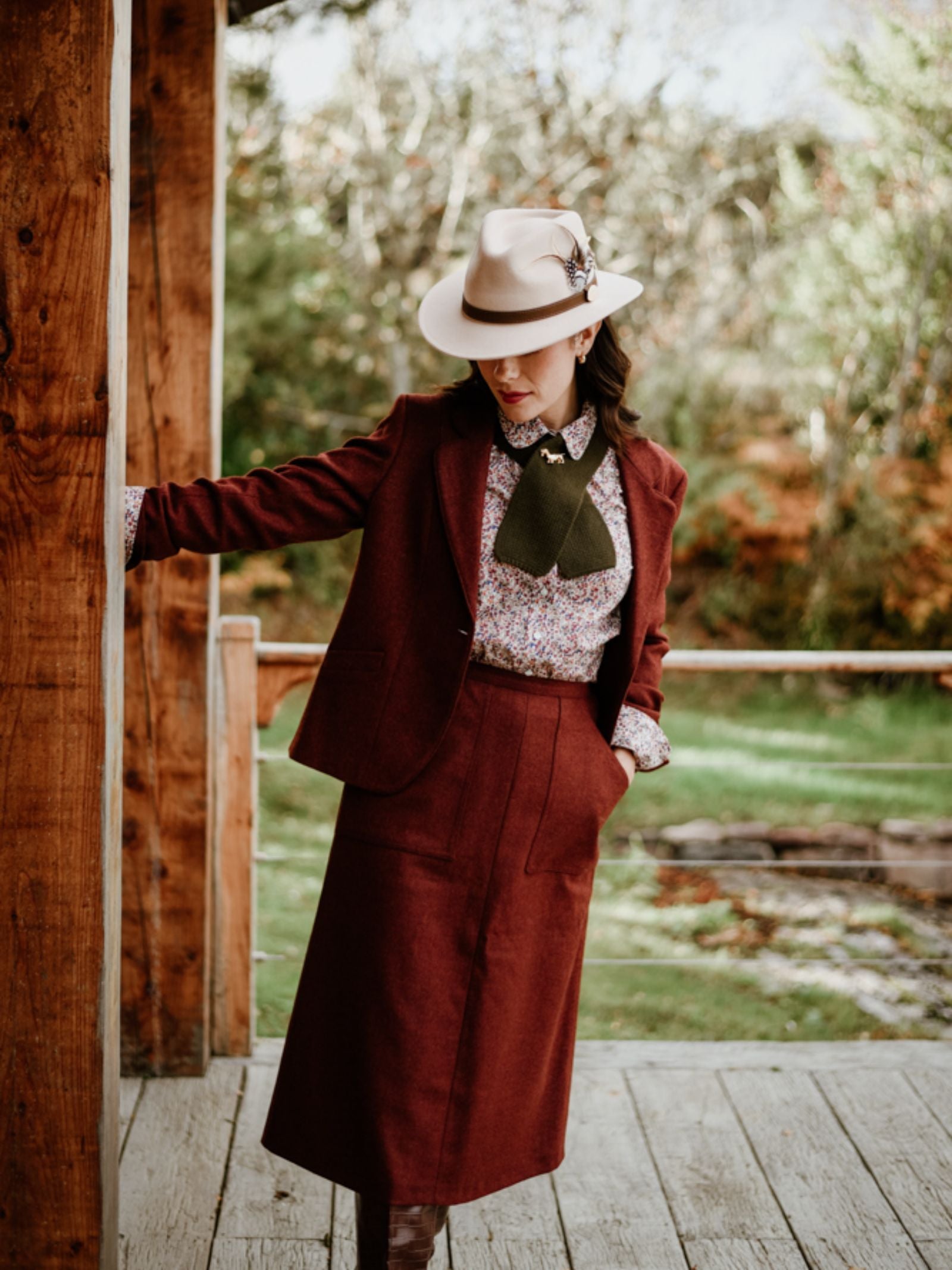 A person in a vintage outfit, wearing the Auld Stock A-Line Skirt from Campbell’s of Beauly, a patterned blouse, green necktie, and beige fedora with a feather, leans against a wooden post on a porch surrounded by greenery.