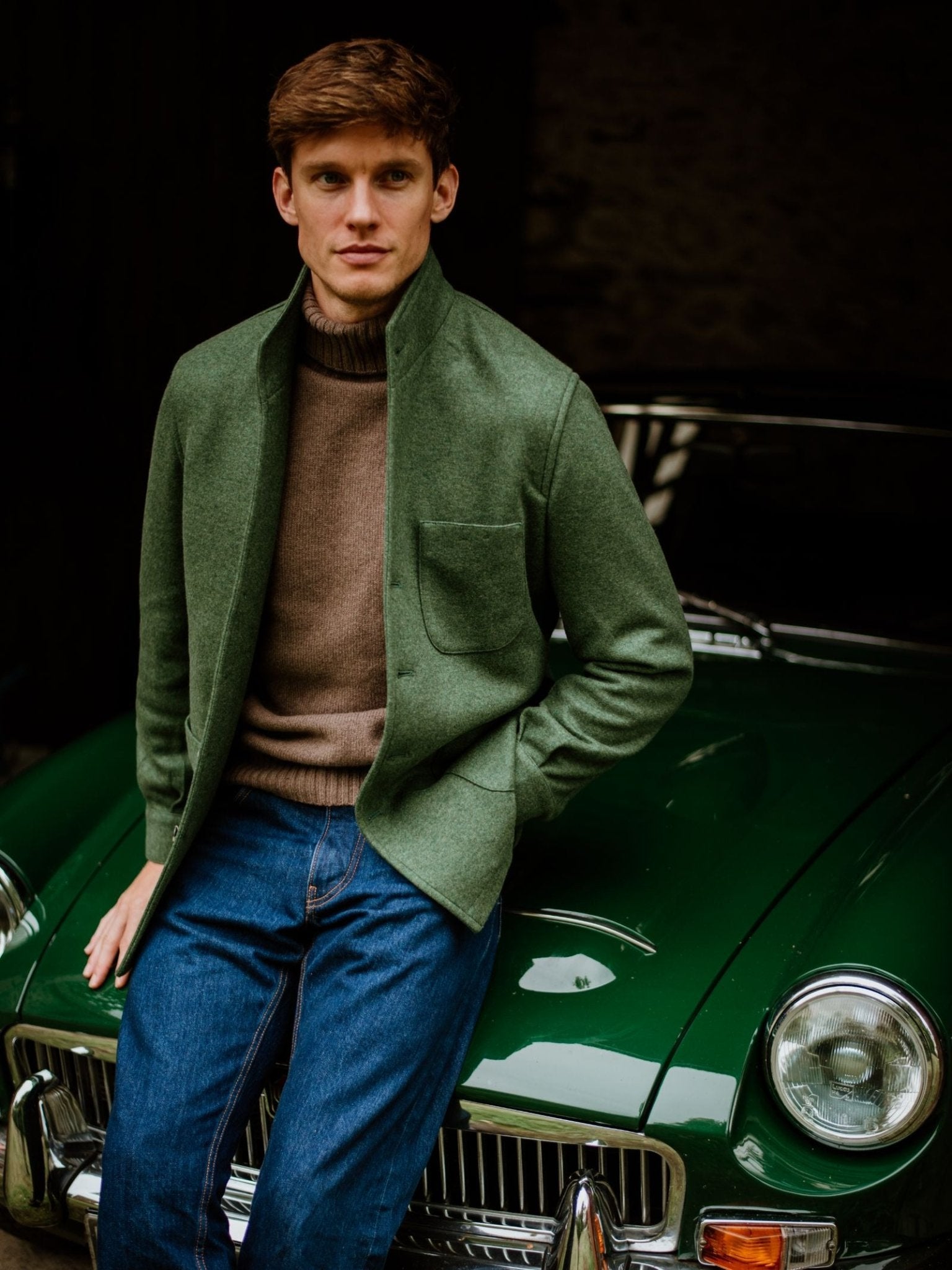 A man in a green jacket, brown turtleneck, and blue jeans leans on a classic green car, his timeless menswear look elevated by the Cashmere Teba Jacket from Campbell's of Beauly, crafted for true luxury.
