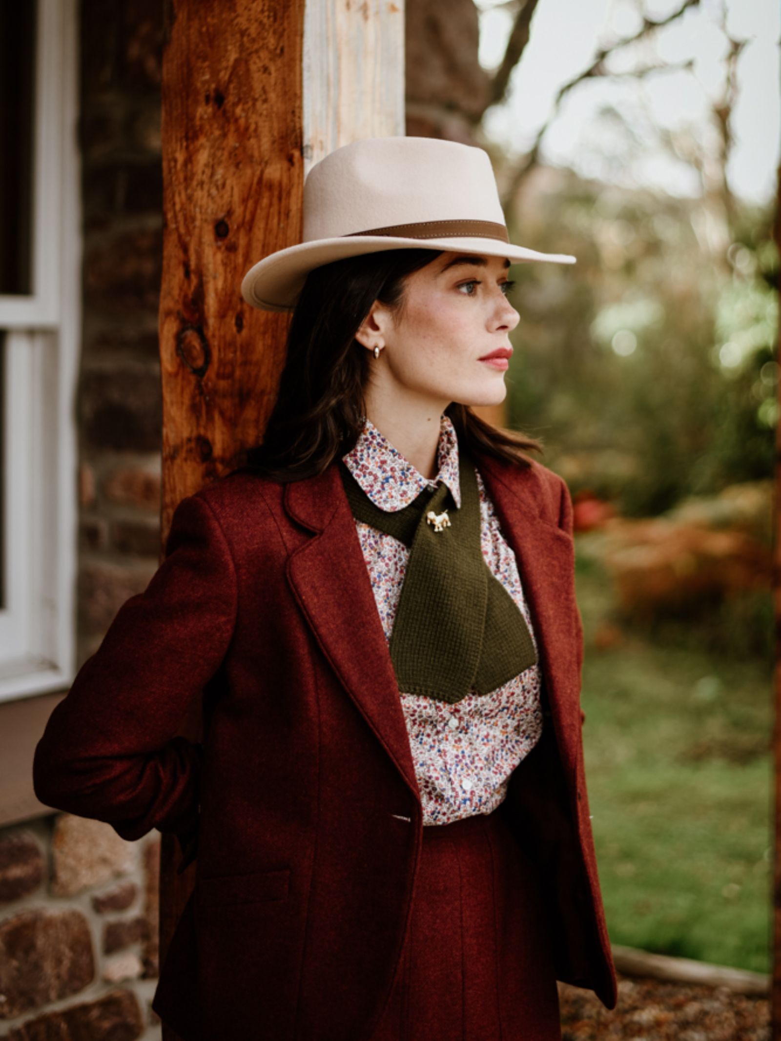 A woman wearing a maroon suit, floral shirt, Campbell’s of Beauly Auld Stock Cashmere Neck Tie in green, and a beige hat stands by a wooden post, looking to the side. The background features greenery and part of a stone building.