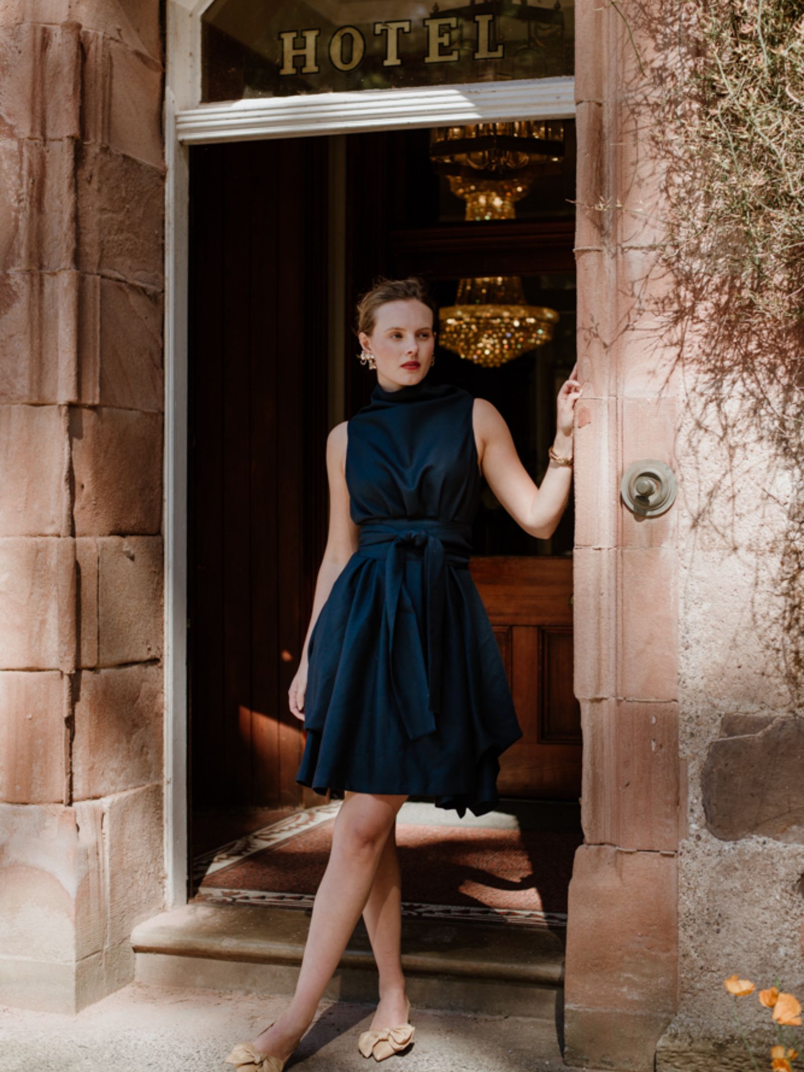 A woman in a sleeveless navy Campbell's of Beauly Reversible Wool Wrap Dress stands in a stone doorway beneath a HOTEL sign, one hand on the frame, gazing outside with chandeliers visible behind her.