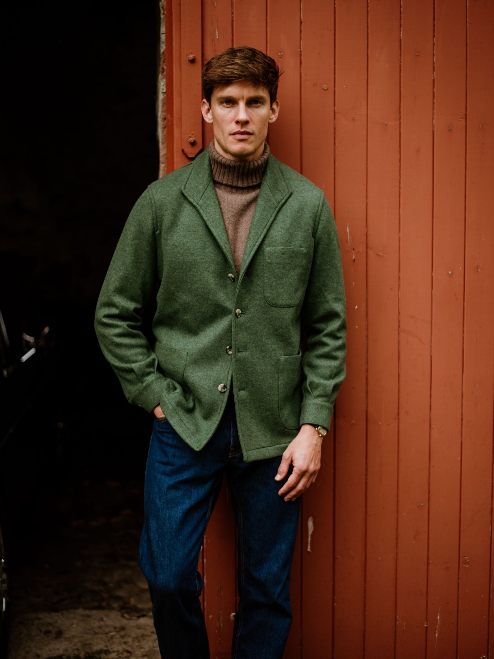 A man in a green Campbell's of Beauly Cashmere Teba Jacket, brown turtleneck, and jeans leans against a rustic red wooden door, showcasing timeless menswear style.