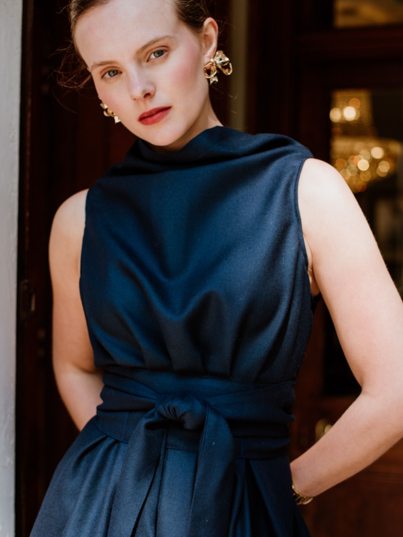 A woman stands indoors wearing a sleeveless, dark navy Reversible Wool Wrap Dress by Campbell's of Beauly. She has light skin, light brown hair pulled back, gold earrings, and is bathed in warm background light.