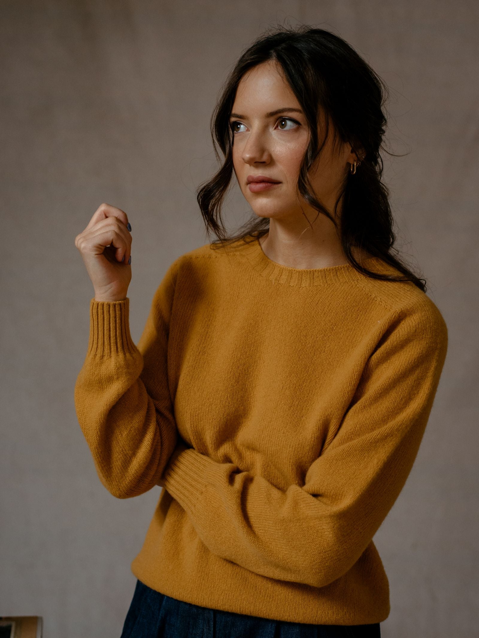 A woman with wavy dark hair wears the Campbell's of Beauly Auld Stock Geelong Lambswool Crew Neck Jumper in mustard yellow and gold hoop earrings, standing against a neutral background with a thoughtful expression.