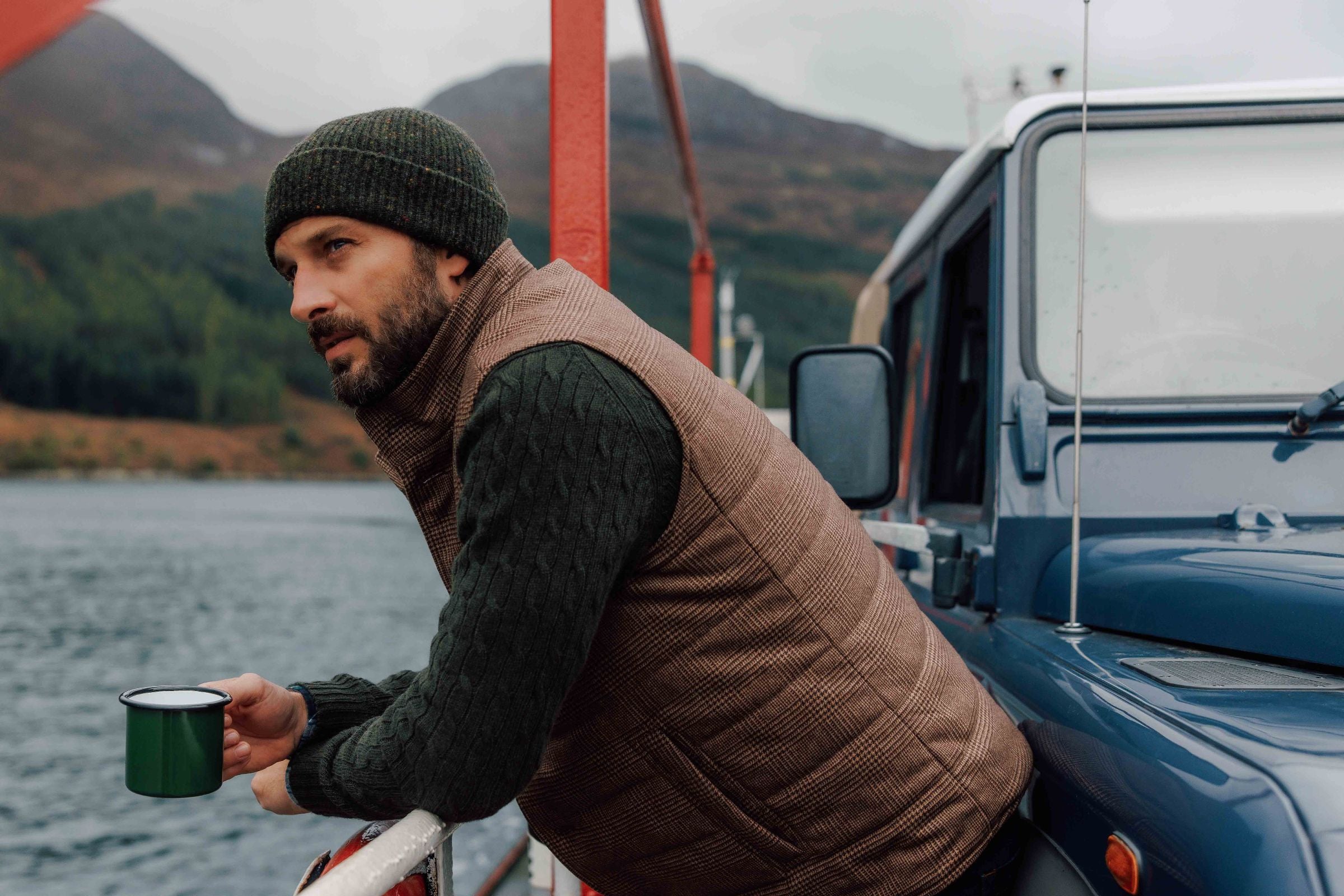 A man in a knit hat and vest leans on a railing beside a blue vehicle, holding a green mug, with a lake and mountains in the background on a cloudy day.