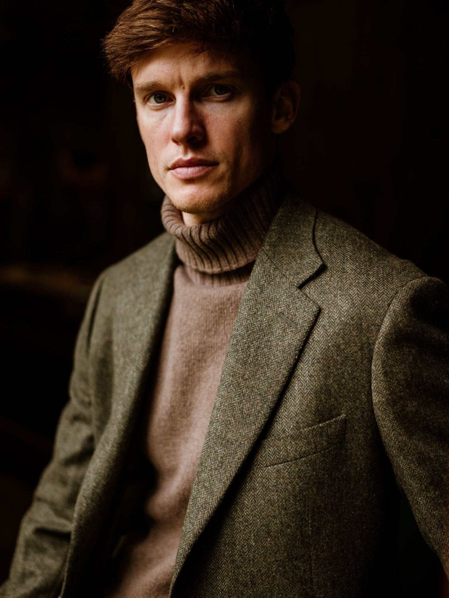 A man with short brown hair wears the Campbell's of Beauly Auld Stock Lambswool Roll Neck sweater and a green tweed blazer, both Scottish-made, as he sits facing the camera against a dark background.