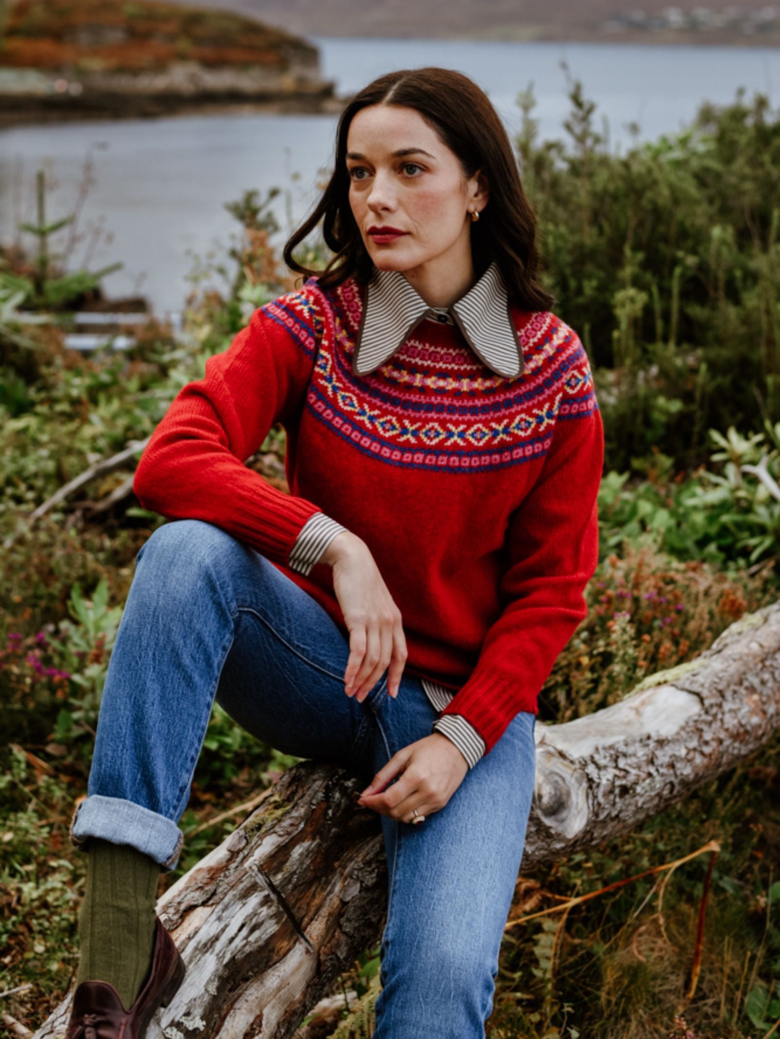 A woman with long dark hair sits on a fallen log outdoors, wearing a red patterned sweater over a striped collared shirt, blue jeans, and brown boots, with greenery and water in the background.