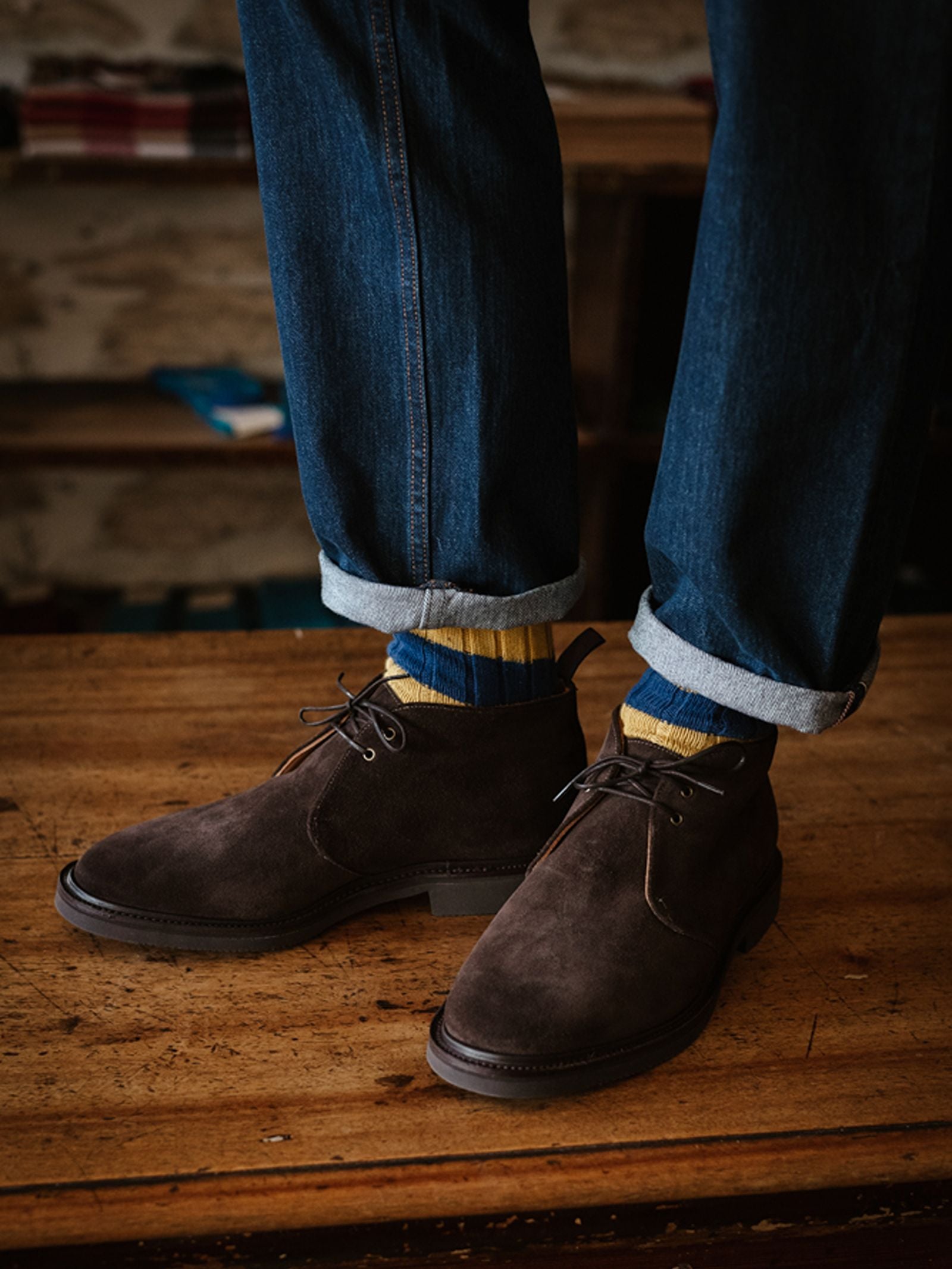A person wearing cuffed blue jeans, yellow and blue striped socks, and Campbell’s of Beauly Lomond Boots in chocolate brown suede with Goodyear welted construction stands on a wooden floor.