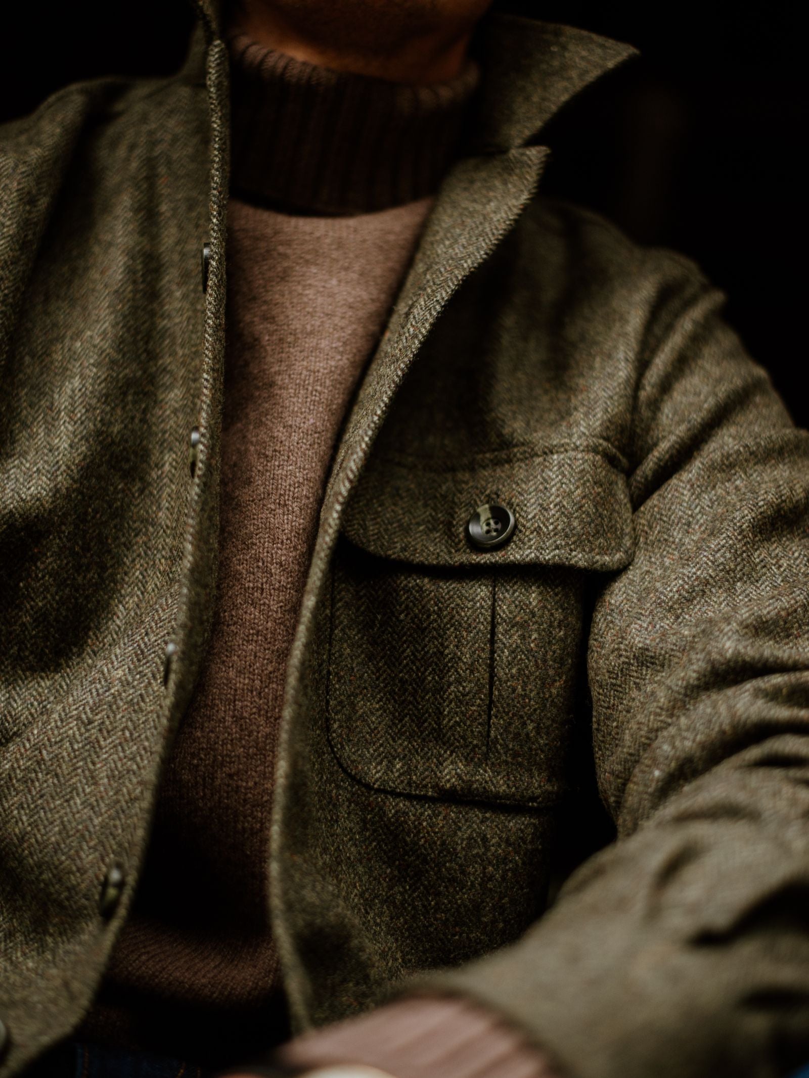 A close-up shows someone in a brown turtleneck layered under the Campbell's of Beauly Casual Tweed Shacket, an olive green British herringbone design with a buttoned chest pocket. Soft light enhances the relaxed tailoring; the face remains unseen.