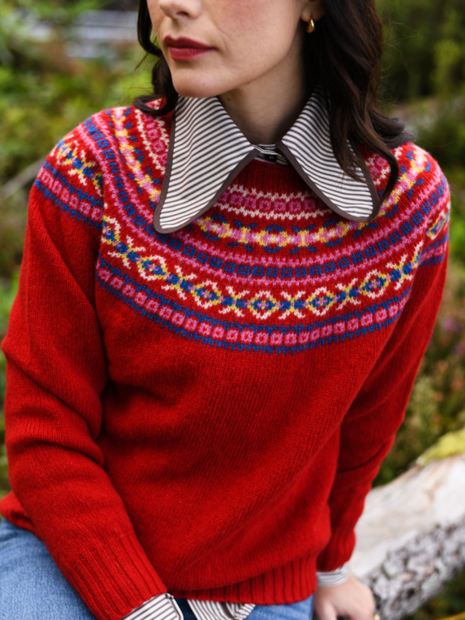 A woman sits outdoors on a log wearing Campbell's of Beauly's Auld Stock Fairisle Crew Jumper, featuring a colorful, intricate pattern over a striped shirt with an oversized collar. The jumper is beautifully knitted in Scotland.