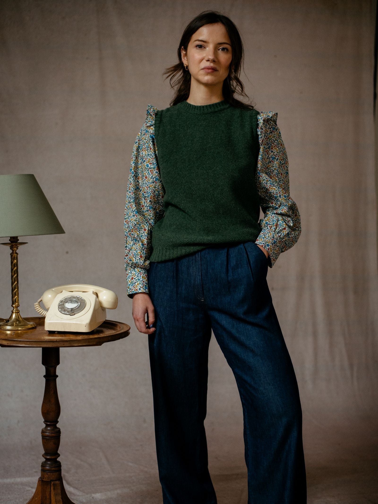 A woman stands with her hand in her pocket, wearing blue jeans, a floral blouse, and the Auld Stock Moss Stitch Crew Tank by Campbell's of Beauly. Beside her is a wooden table with a green lamp and beige rotary phone against a neutral backdrop.
