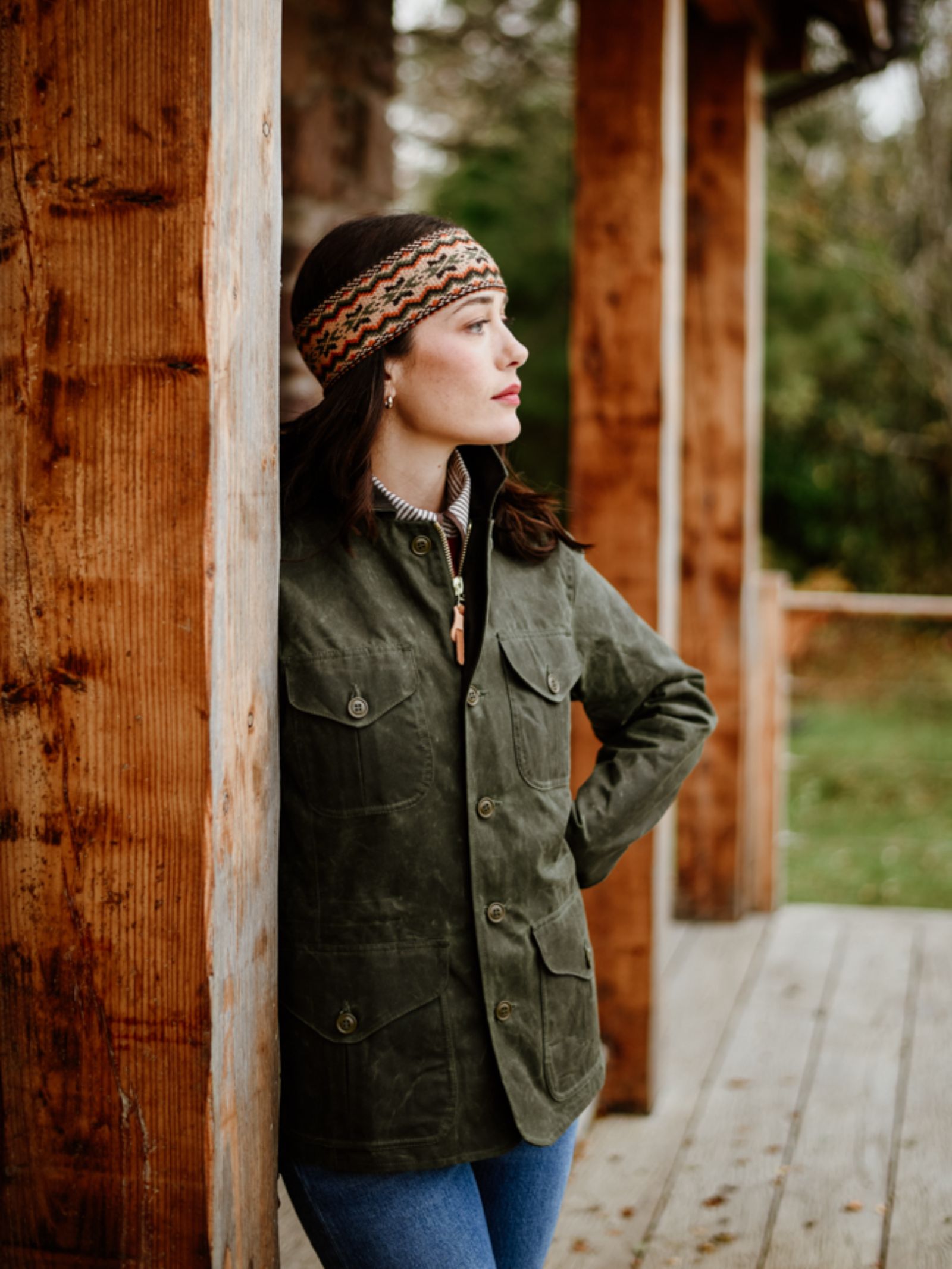 A woman wearing a Campbell's of Beauly Fairisle Headband and a green jacket leans against a porch post, hands behind her back, gazing sideways. A blurred green backdrop evokes the serene charm of the Scottish Borders.