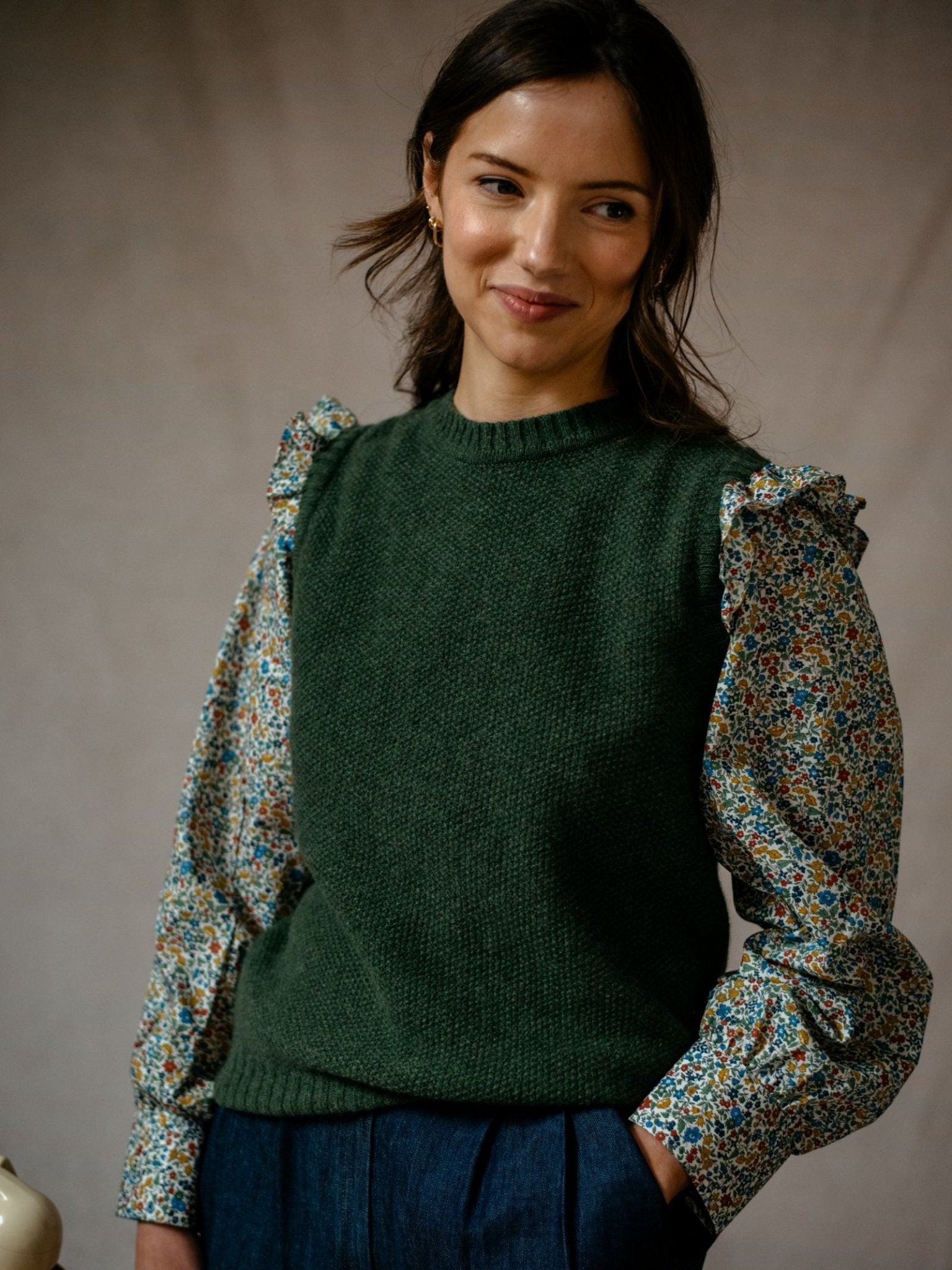A woman with dark hair smiles, wearing Campbell's of Beauly's Auld Stock Liberty Print Peplum Shirt with ruffled shoulders under a green sleeveless knit sweater and dark blue pants, standing against a neutral backdrop.