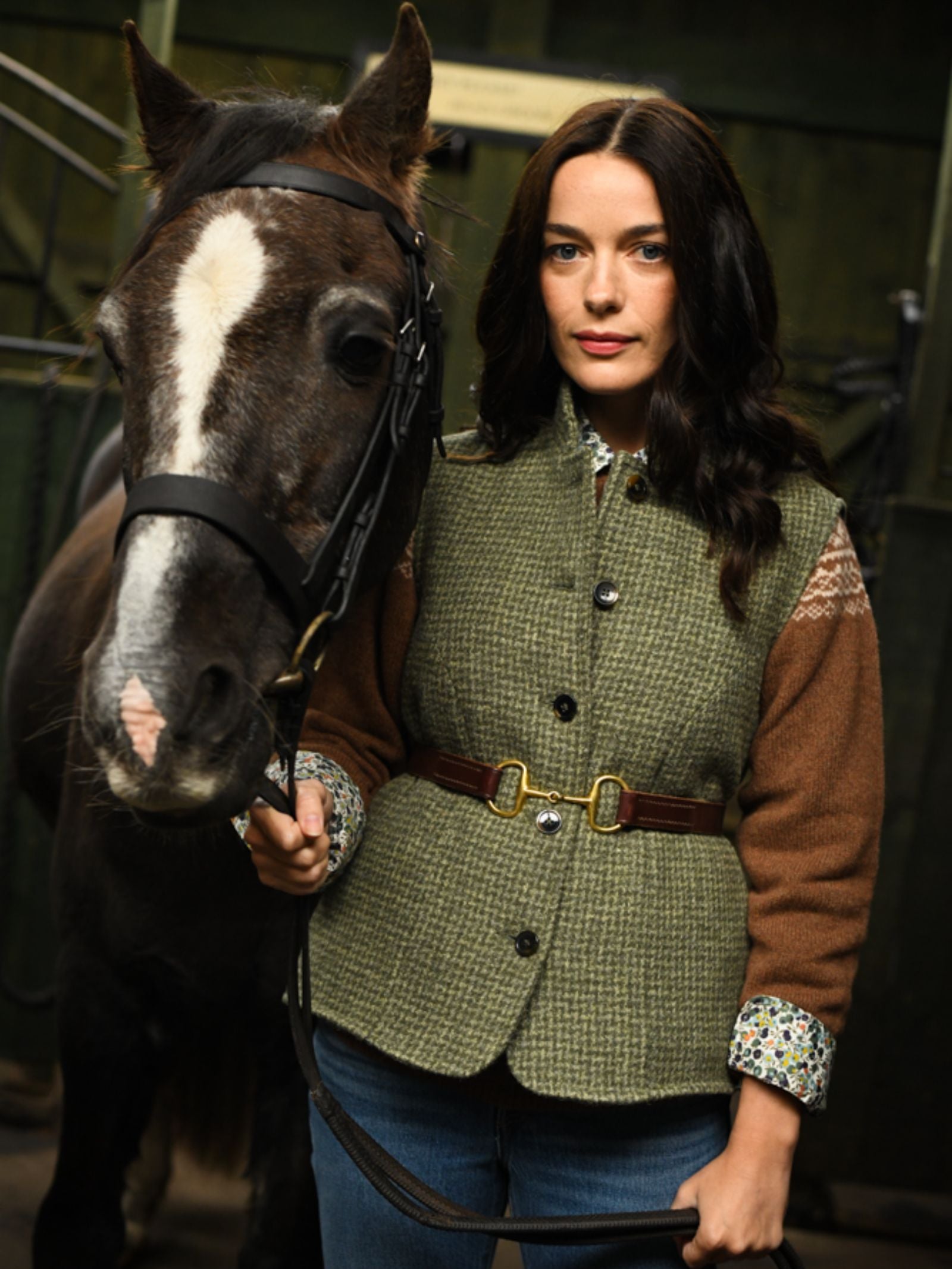 A woman with long dark hair wears the Campbell's of Beauly Auld Stock Harris Tweed Houndstooth Gilet as she stands in a stable, holding the bridle of a dark brown horse with a white stripe on its face.
